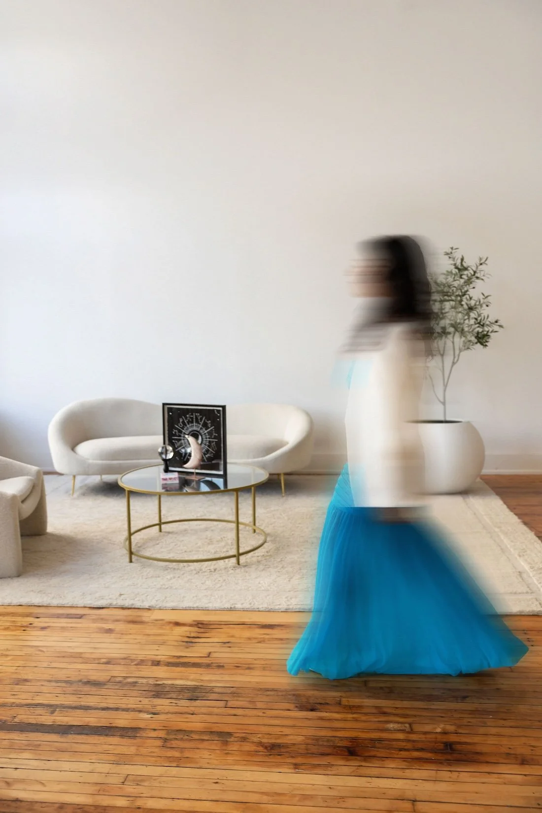 A blurred woman in a white top and blue skirt walking past a modern living room with a white sofa, a round glass coffee table with gold trim, a black decorative clock, and a large ceramic pot with a plant.