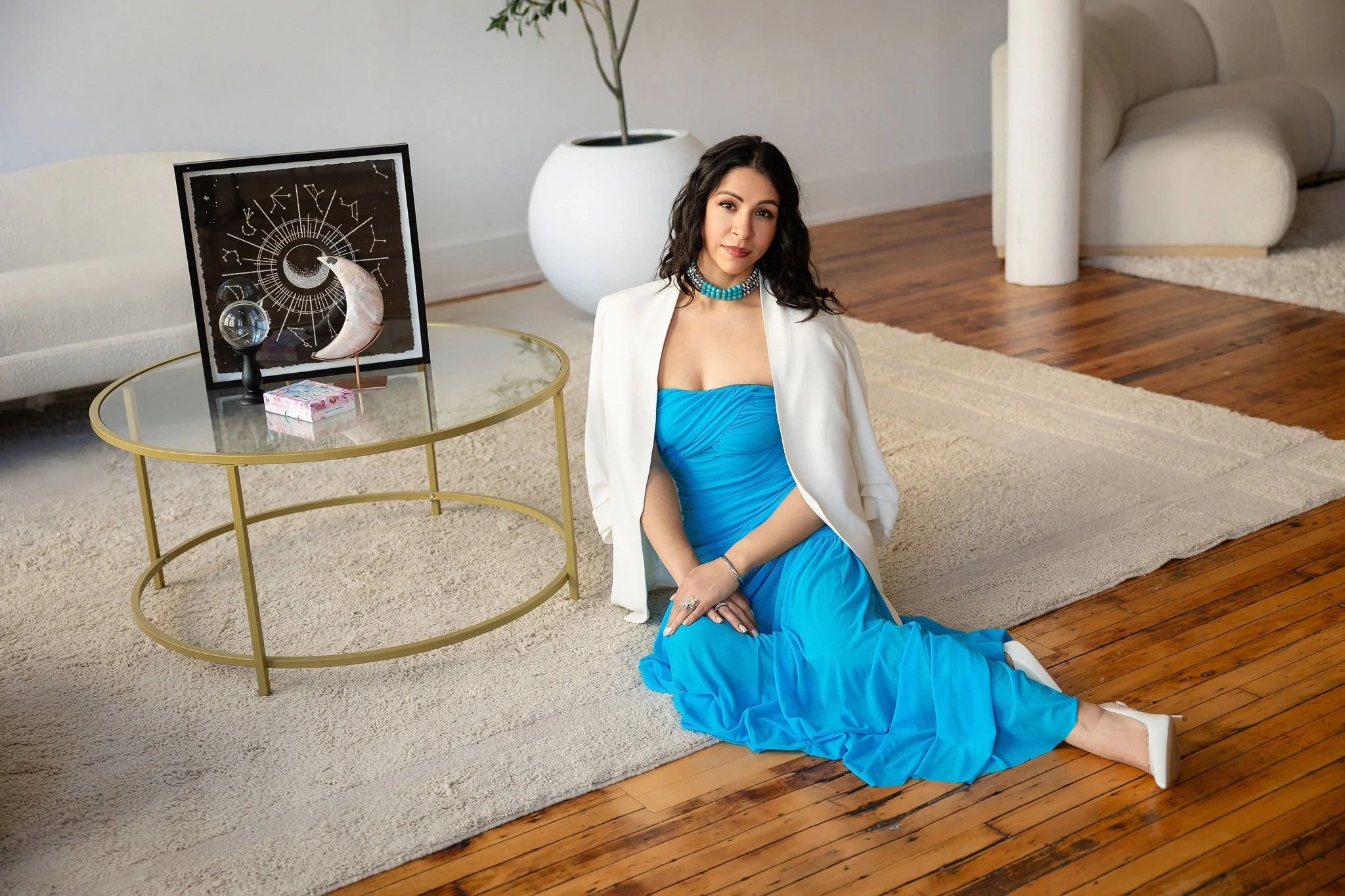 A woman dressed in a blue gown with white blazer and turquoise necklace, sitting on a rug in a modern living room with wooden floors. A black framed celestial art piece and a white vase with a plant are on a glass-topped table nearby.