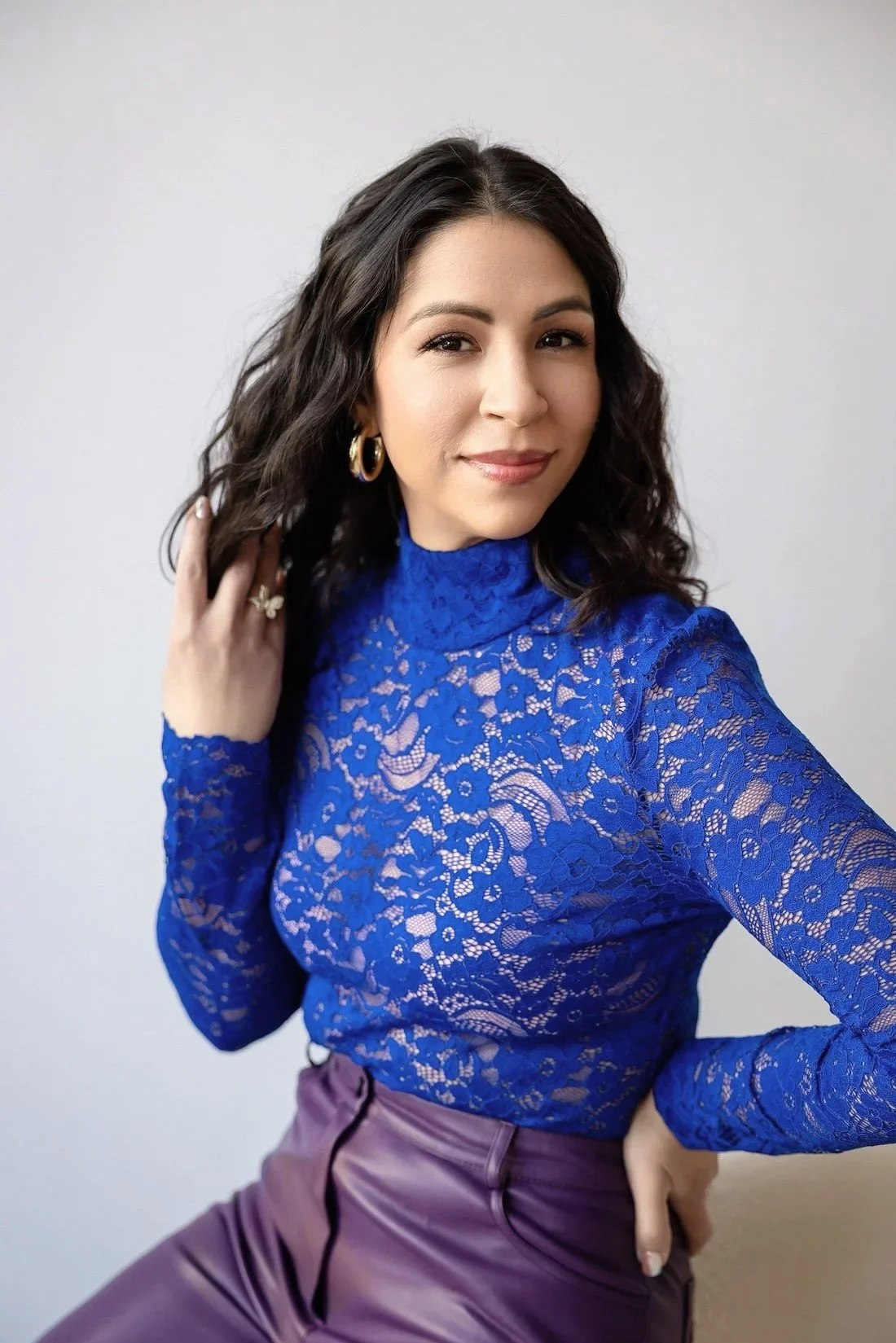 A woman taking a selfie wearing a bright blue lace top and purple pants, with dark wavy hair, gold hoop earrings, and a ring, against a plain white background.