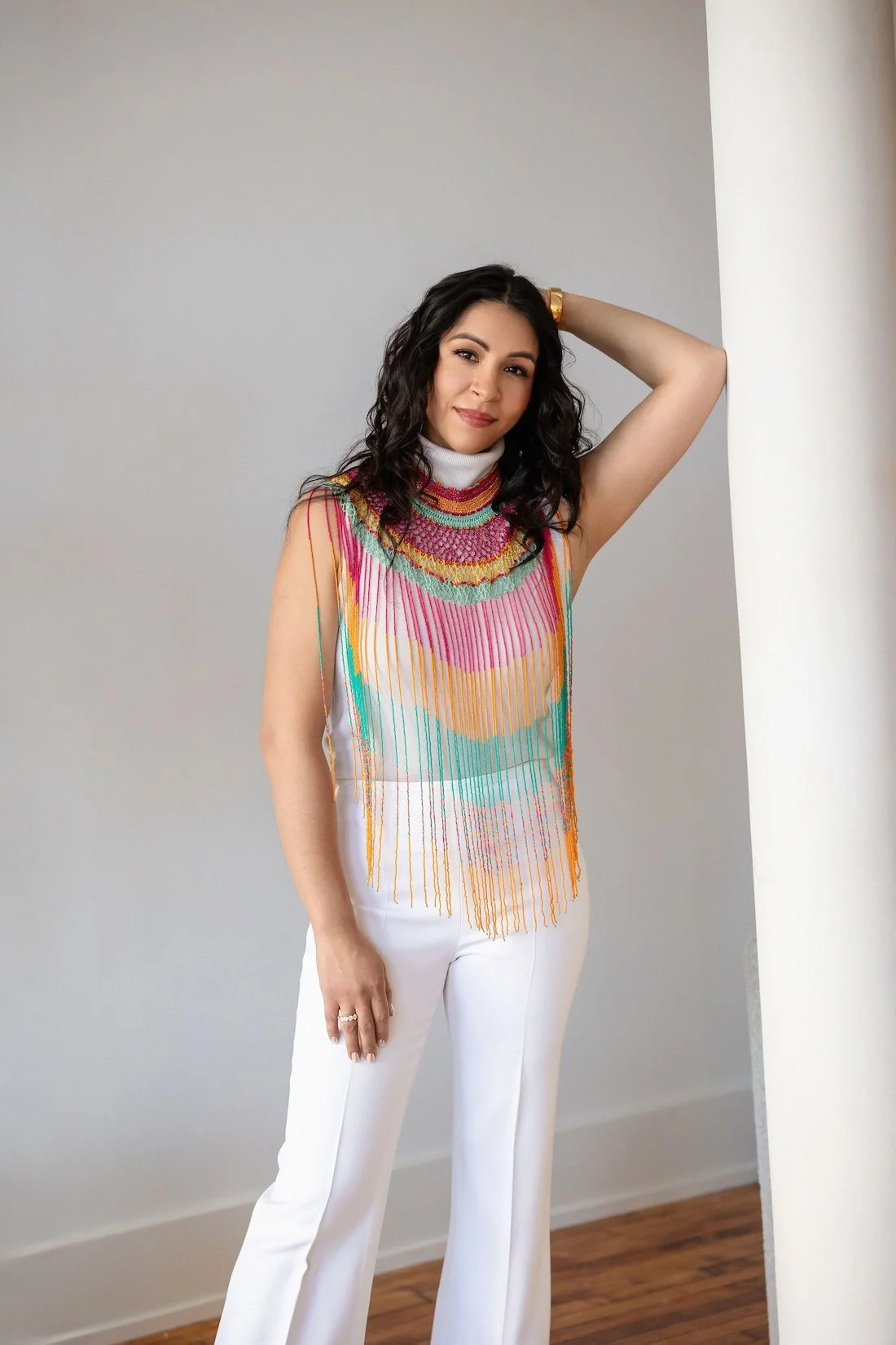 A woman with black wavy hair wearing a sleeveless, colorful, embroidered top and white pants, standing against a plain wall.