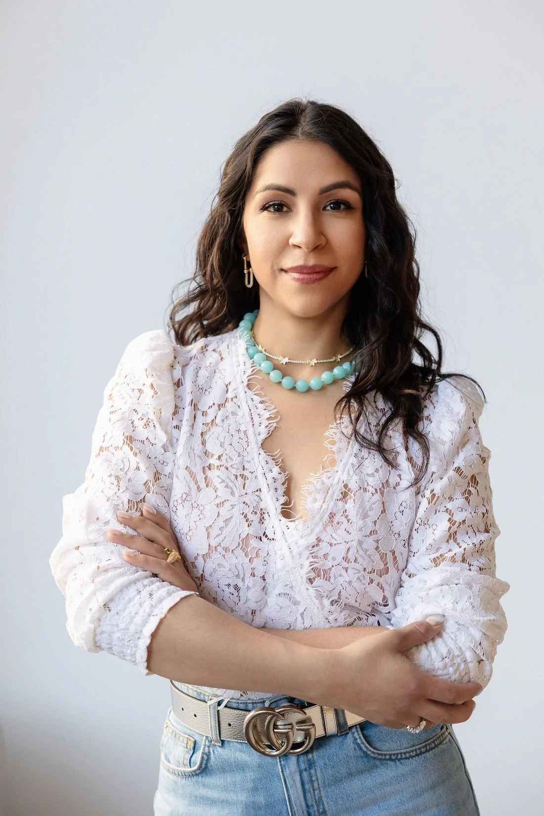 A woman with dark curly hair and light skin wearing a white lace top, turquoise beaded necklace, and gold jewelry, standing against a plain white background.