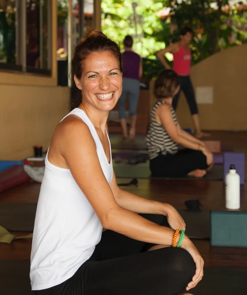 Eine Frau sitzt in einer Yoga-Klasse, lächelnd, mit dunklem Haar in einem Dutt, tragt ein weißes Tanktop und schwarze Hosen, im Hintergrund andere Frauen, Yoga-Matten und eine mit Wasser gefüllte Flasche.