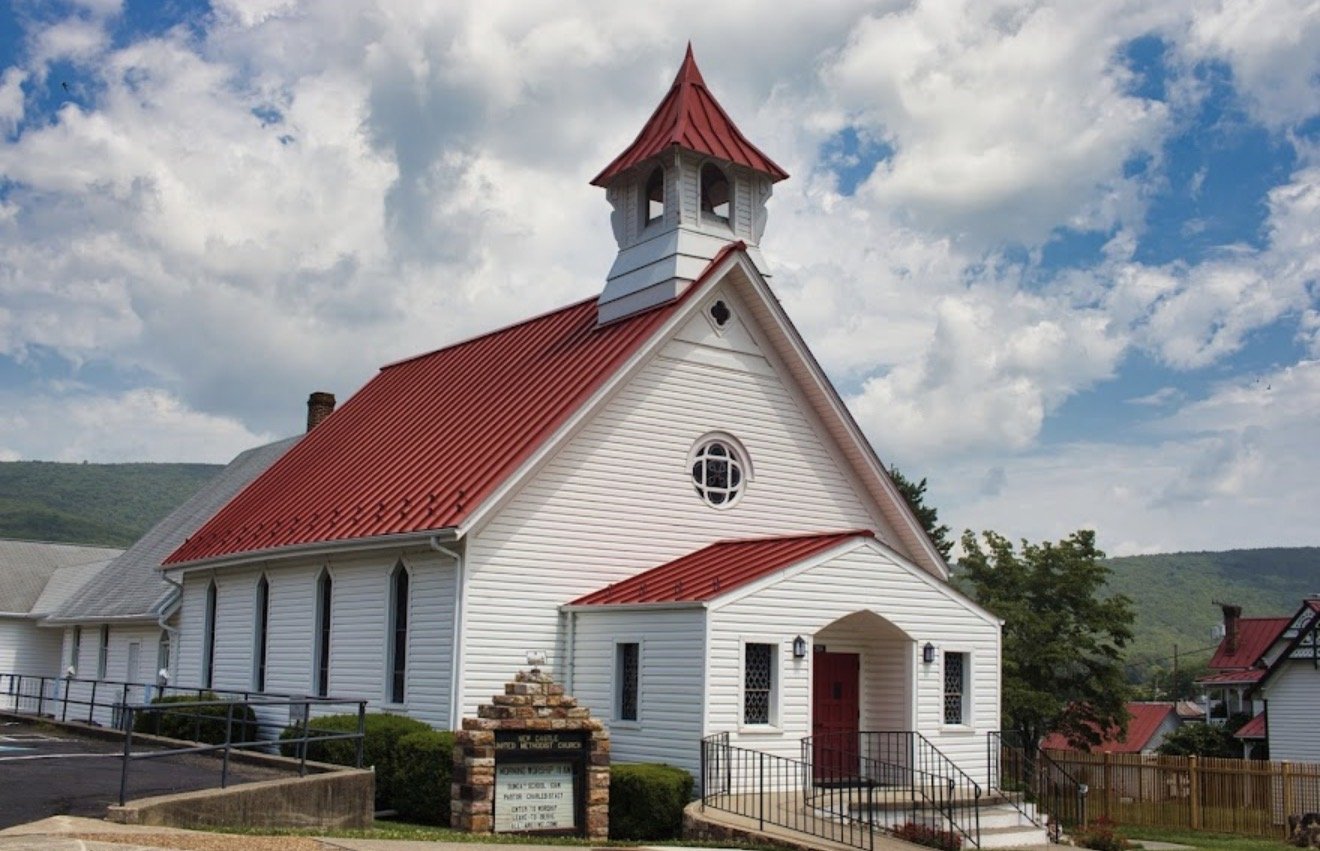 White church with red roof and steeple, situated in a small town, surrounded by hills and neighboring buildings, under a partly cloudy sky.