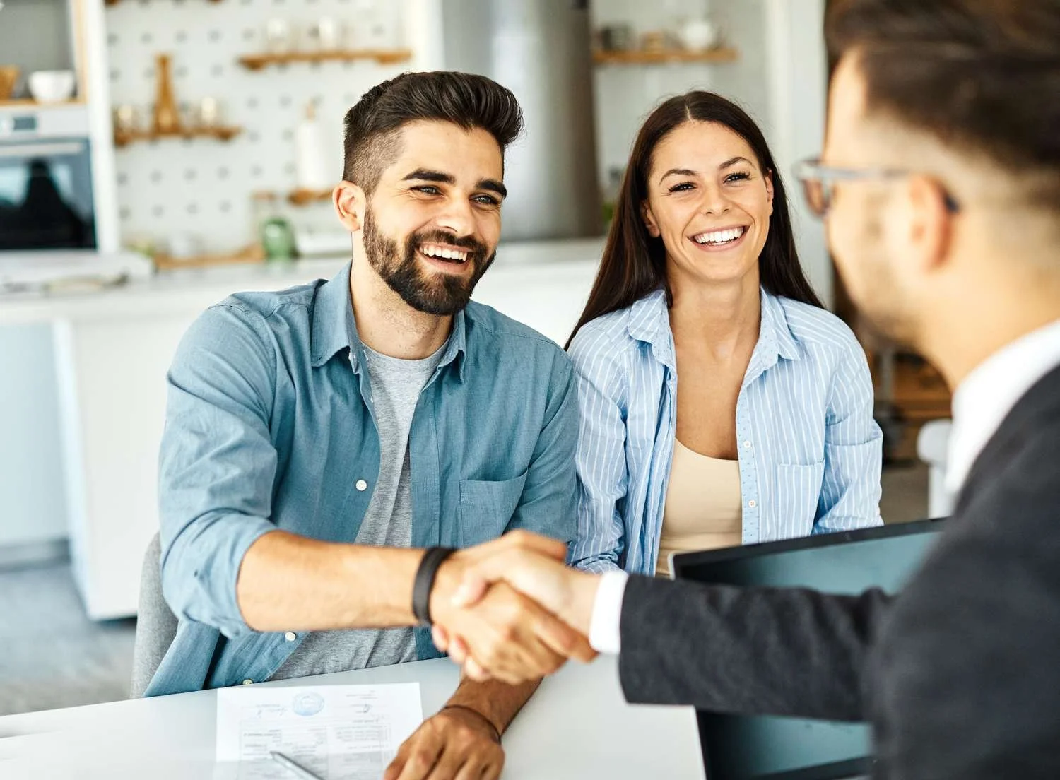 Three people in a professional setting shaking hands and smiling, with a woman and man facing a person with glasses in the foreground.