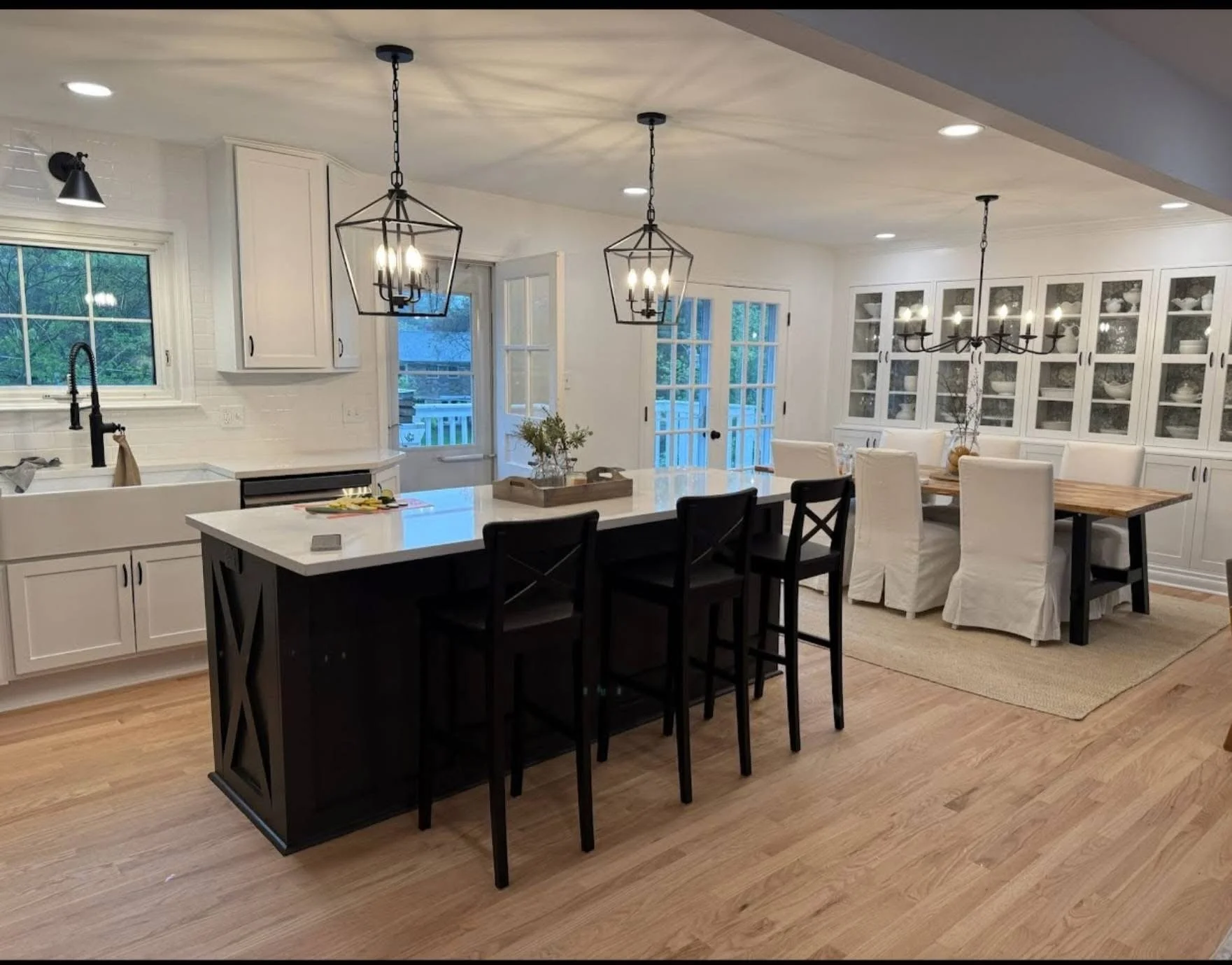 Open-concept kitchen and dining area with white cabinetry, large island with black base, black and white seating, glass-front cabinets displaying dishes, chandelier, and pendant lights, with hardwood flooring and large windows.
