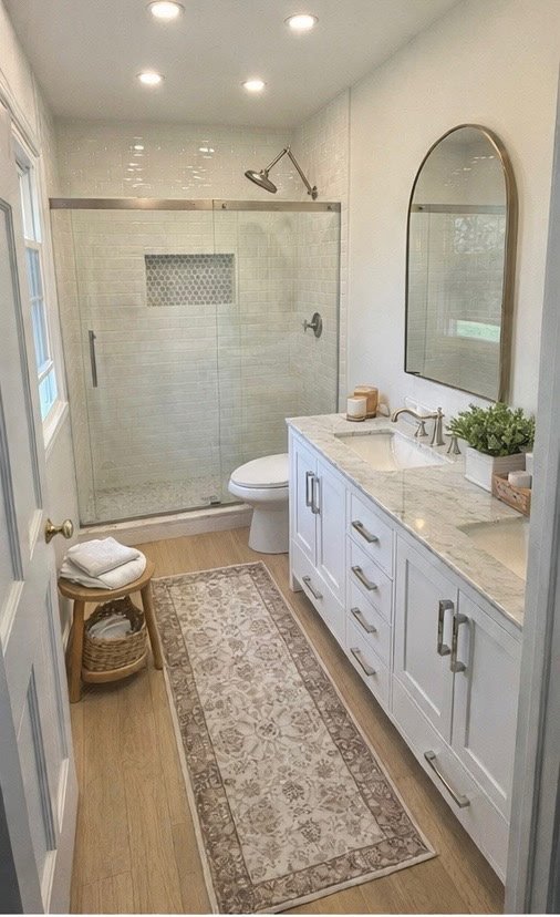 Bathroom with a glass-enclosed shower, white vanity with marble countertop, oval mirror, potted plant, and a small wooden stool with towels. Wooden flooring and a decorative rug.