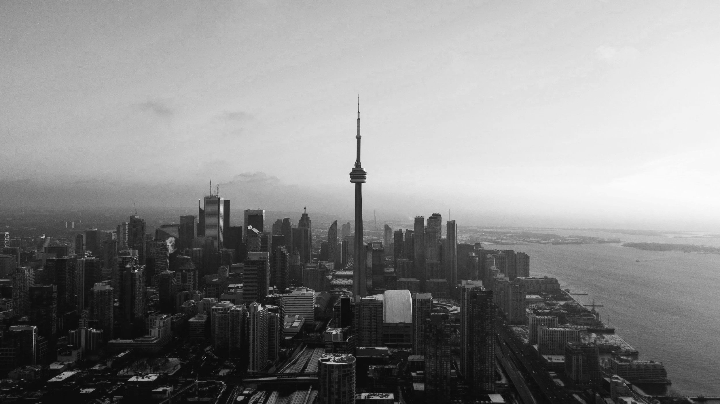Black and white aerial view of Toronto skyline with CN Tower in the center, showing numerous tall skyscrapers and the waterfront in the background.