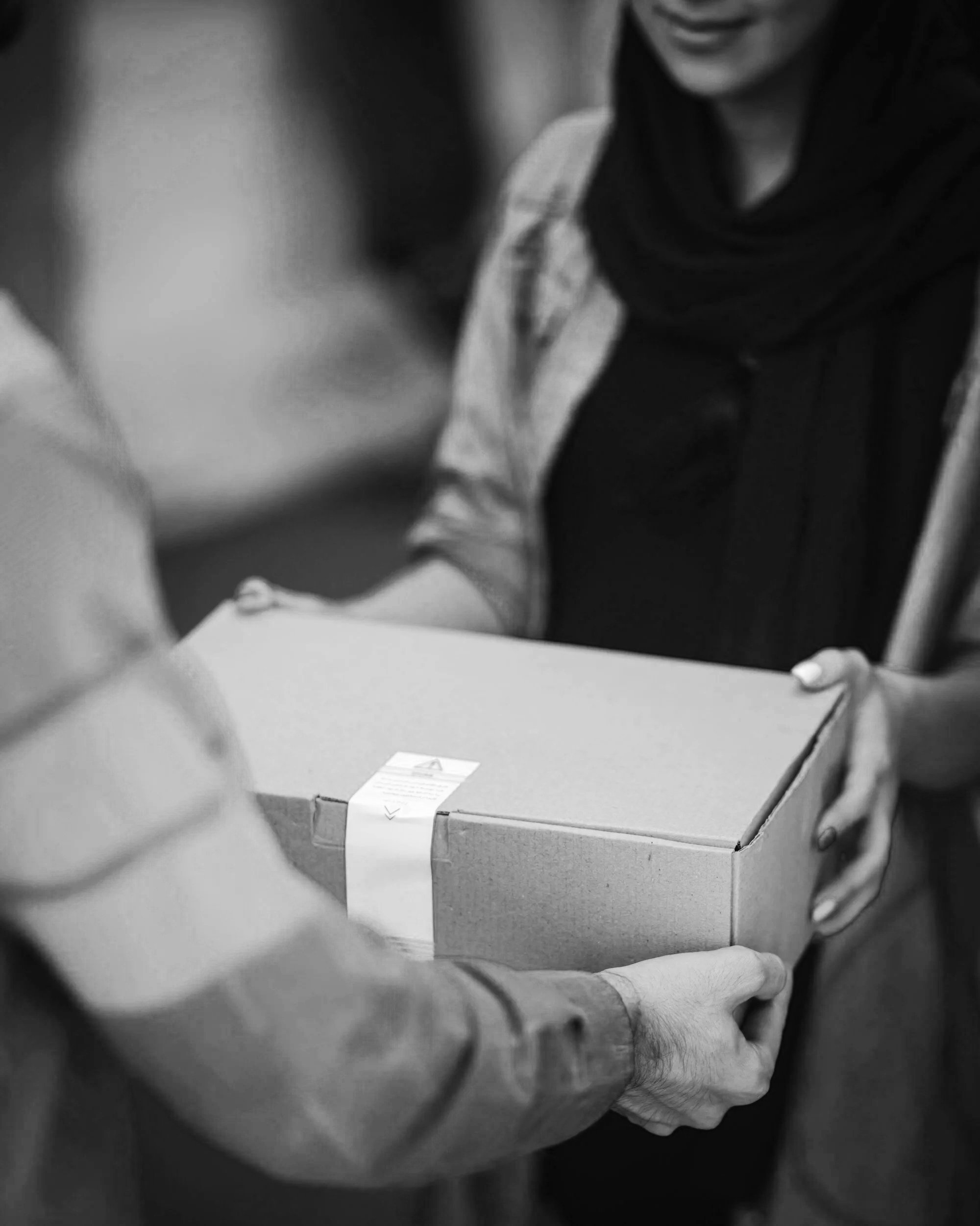 Close-up of two individuals passing a cardboard box, one person in the foreground and other in the background, both wearing casual clothing and a scarf.