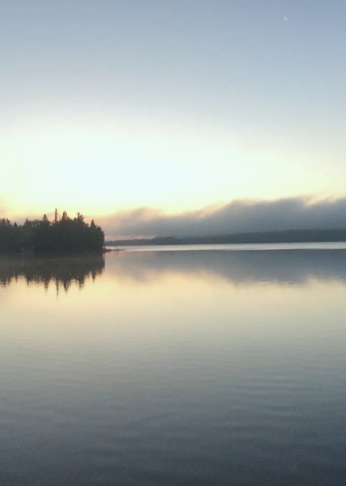A calm body of water reflecting the sky, with trees on the left side and a foggy, cloudy horizon in the distance at sunrise or sunset.