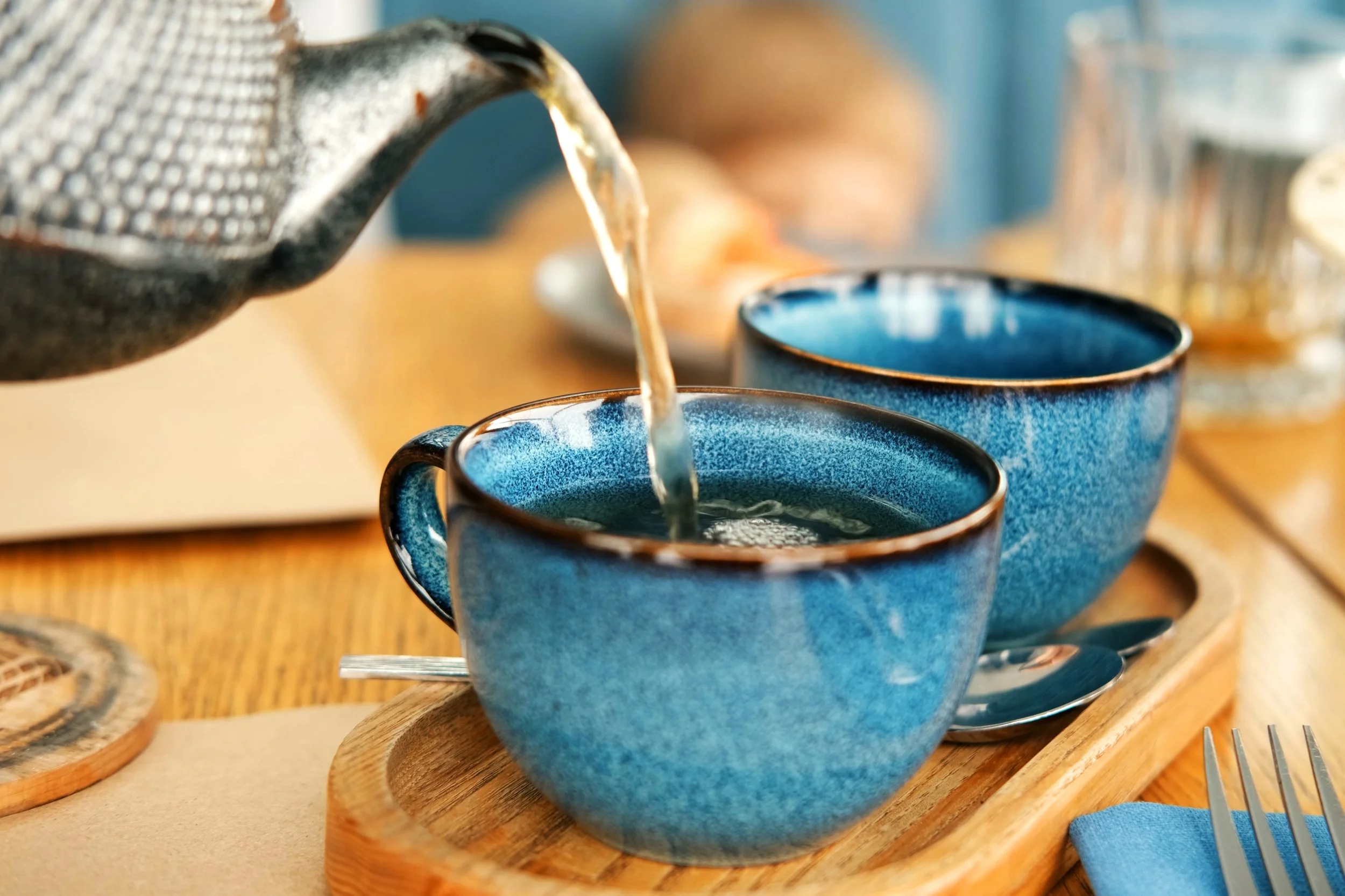 Pouring coffee into blue ceramic mugs on a wooden tray at a breakfast table.