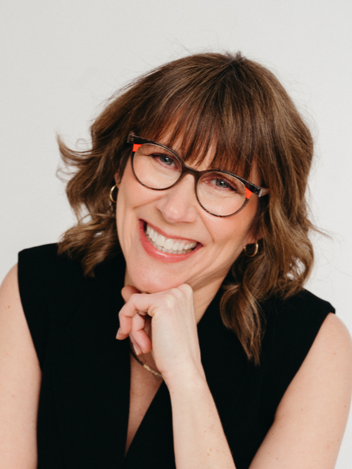 A woman with shoulder-length brown hair, glasses, and earrings, smiling and resting her chin on her hand, against a plain white background.