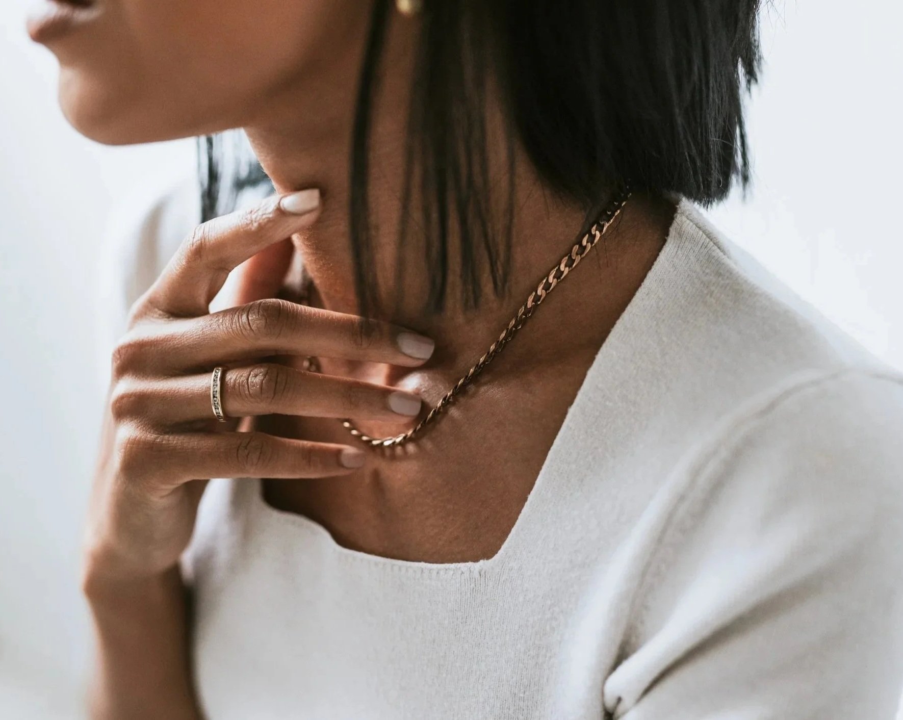 A woman with short dark hair wearing a white top, a gold chain necklace, and a silver ring, gently touching her neck with her fingers.