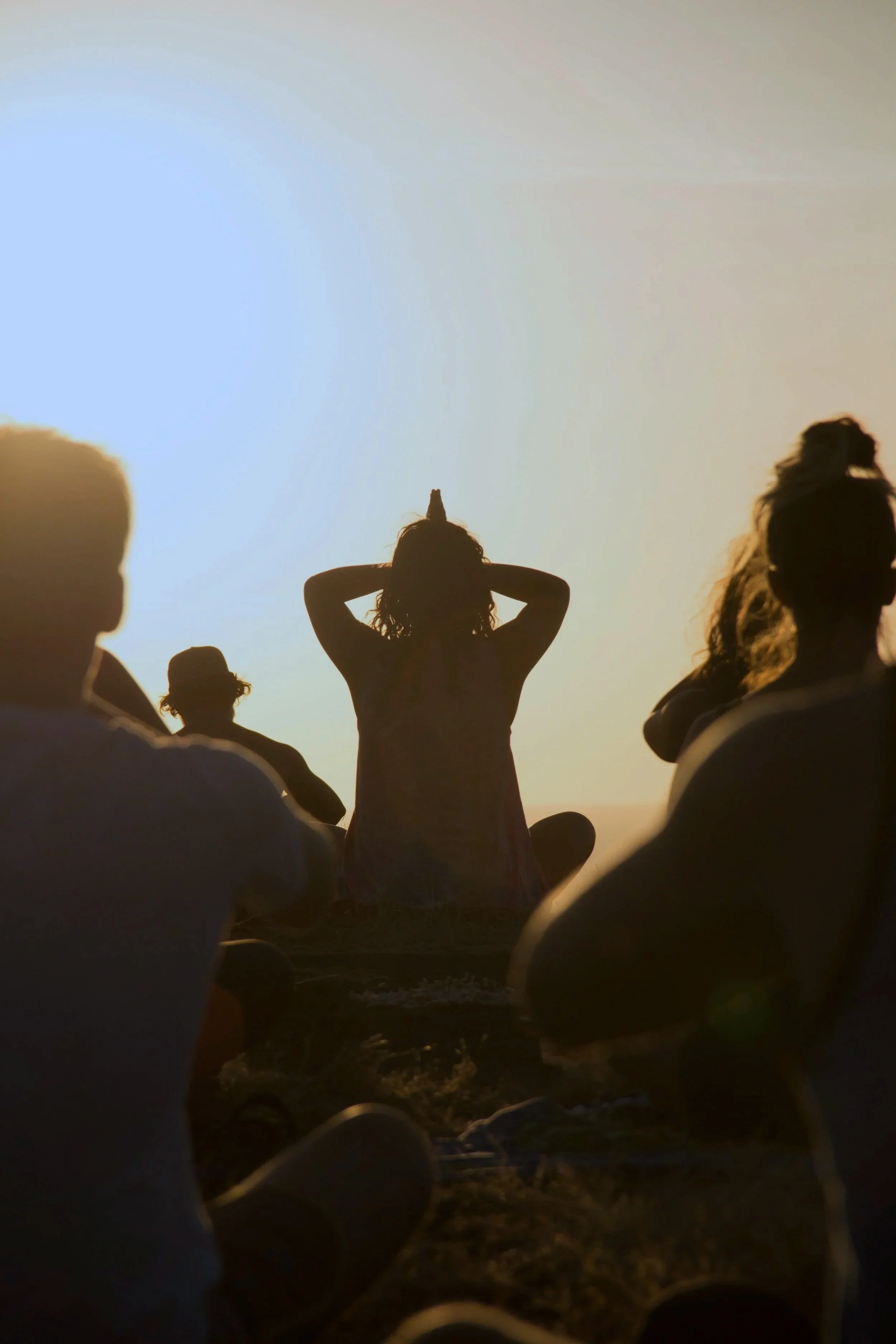 Silhouette of people sitting and standing on a beach during sunset, with one woman standing and holding her arms behind her head, sky glowing with warm colors.