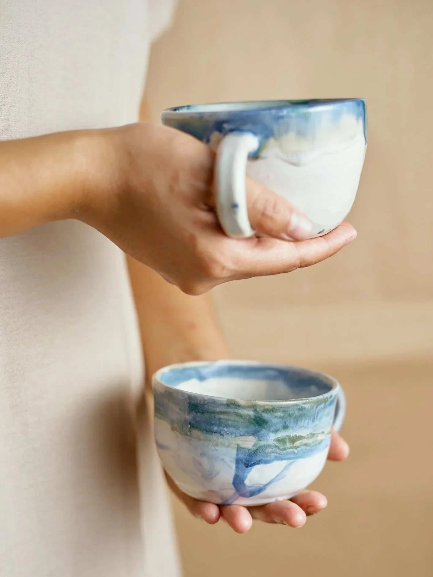 A person holding a blue and white ceramic cup with both hands, with another similar cup below also held in one hand, against a beige background.