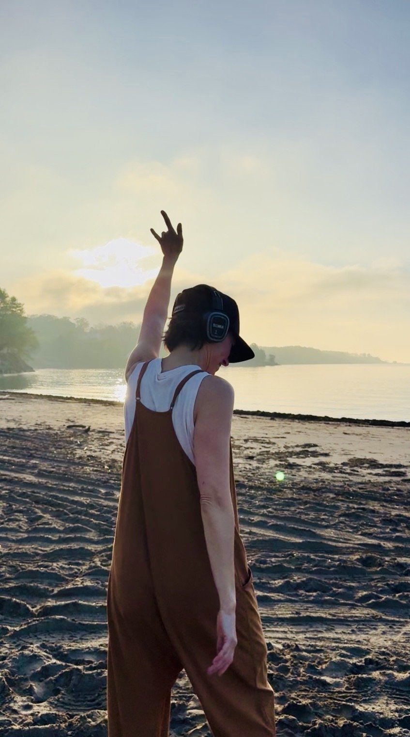 A woman on the beach at sunset, wearing headphones, making a peace sign with her right hand, and looking down.