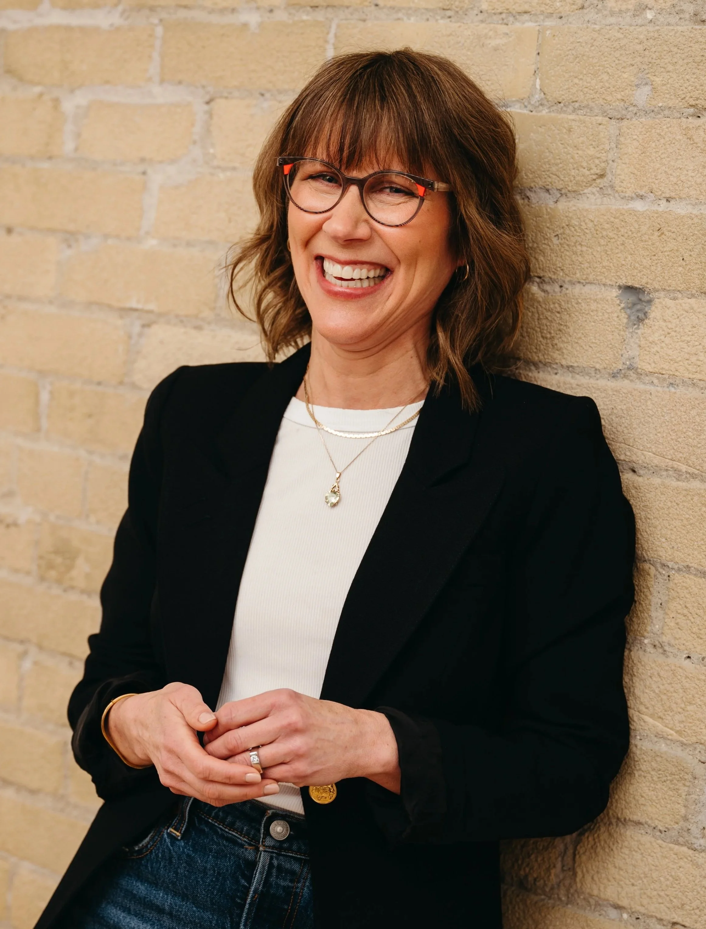 A woman with shoulder-length brown hair, glasses, and a wide smile, standing against a beige brick wall. She is wearing a white top, black blazer, and jewelry, with her hands clasped.