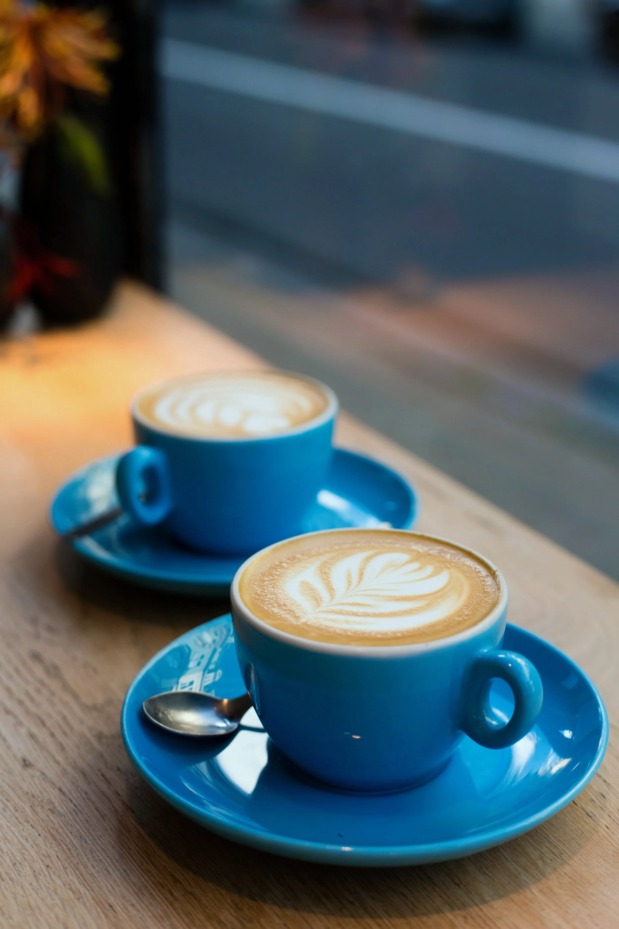 Two blue cups of coffee on matching blue saucers with stainless steel teaspoons, placed on a wood table, with latte art on top of each coffee.