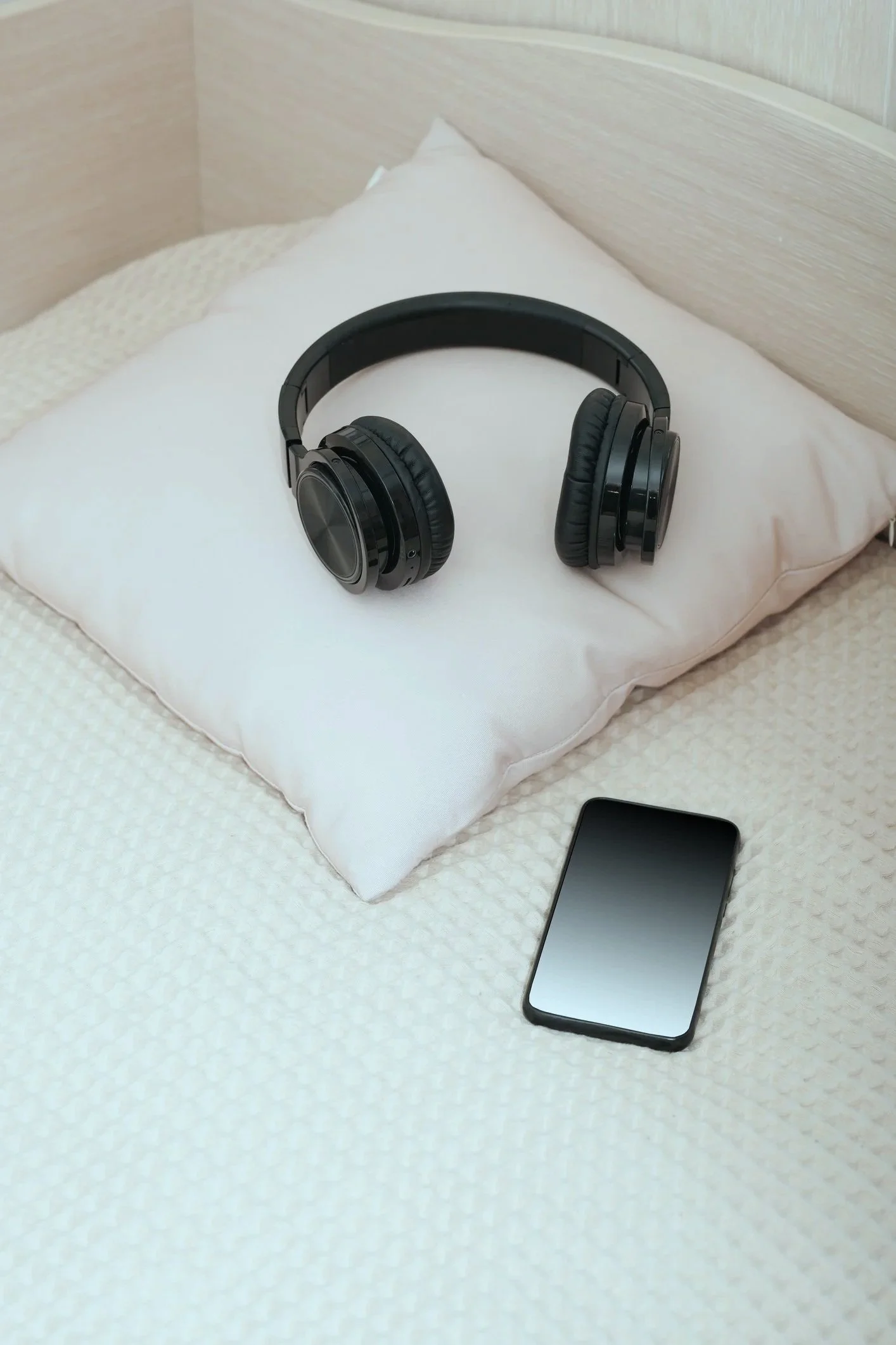 A pair of black over-ear headphones resting on a white pillow on a bed, with a smartphone lying nearby on a textured white bedsheet.