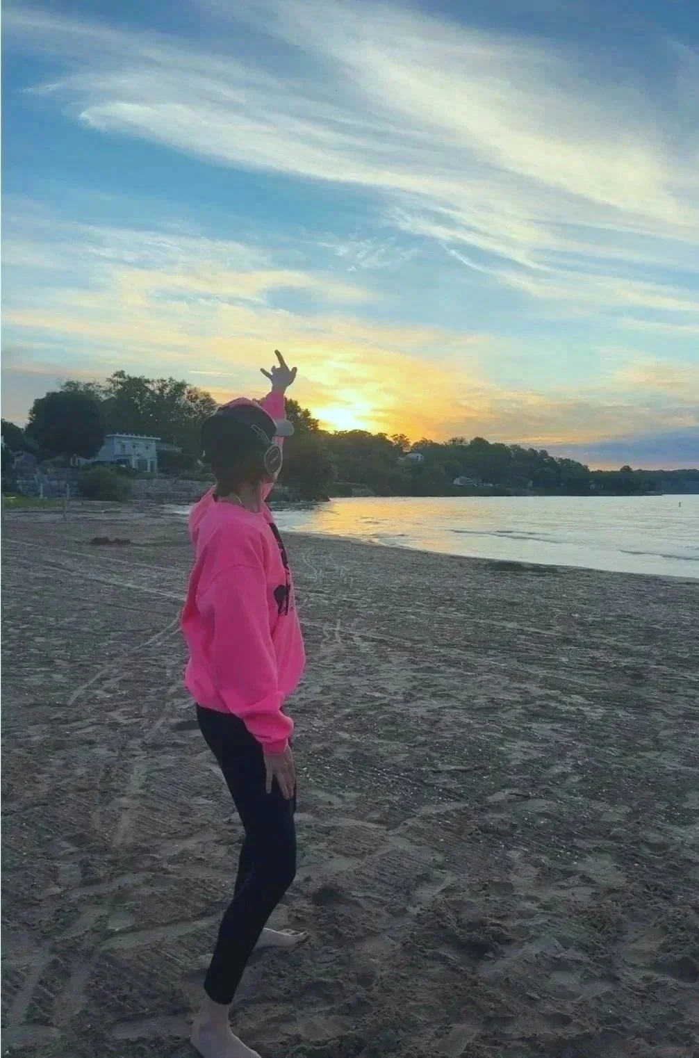 A person standing on the beach wearing a pink hoodie and headphones, reaching up with one hand towards the sunset over the water, with a cloudy sky and trees in the background.