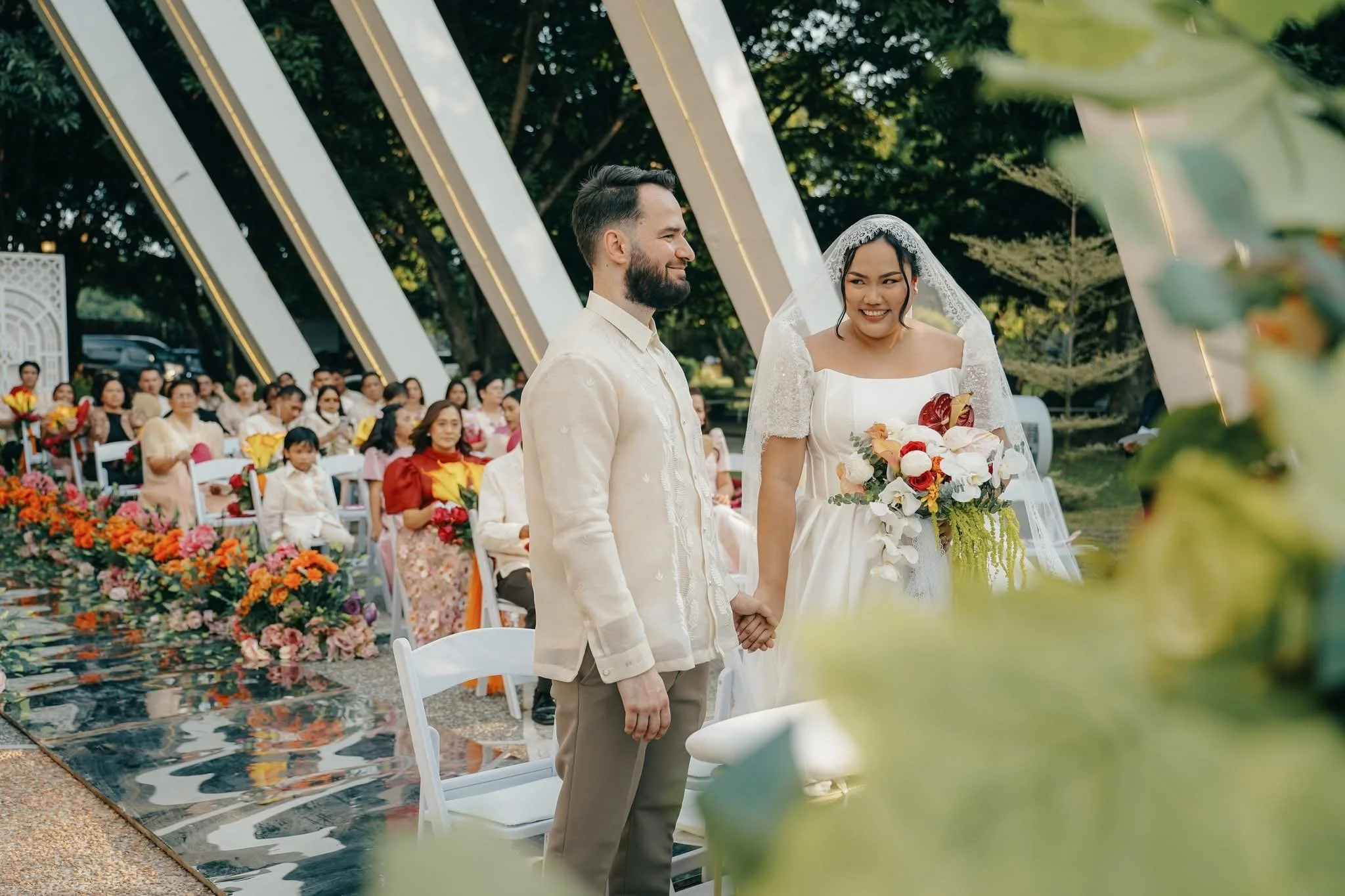 A wedding ceremony outdoors with a bride and groom holding hands and surrounded by seated guests and colorful flowers.