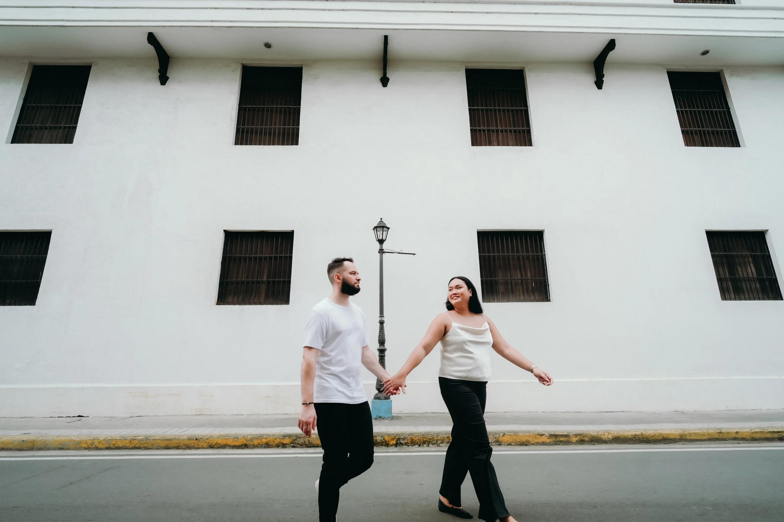 A man and woman holding hands walking across the street in front of a white building with black windows and a black streetlamp.