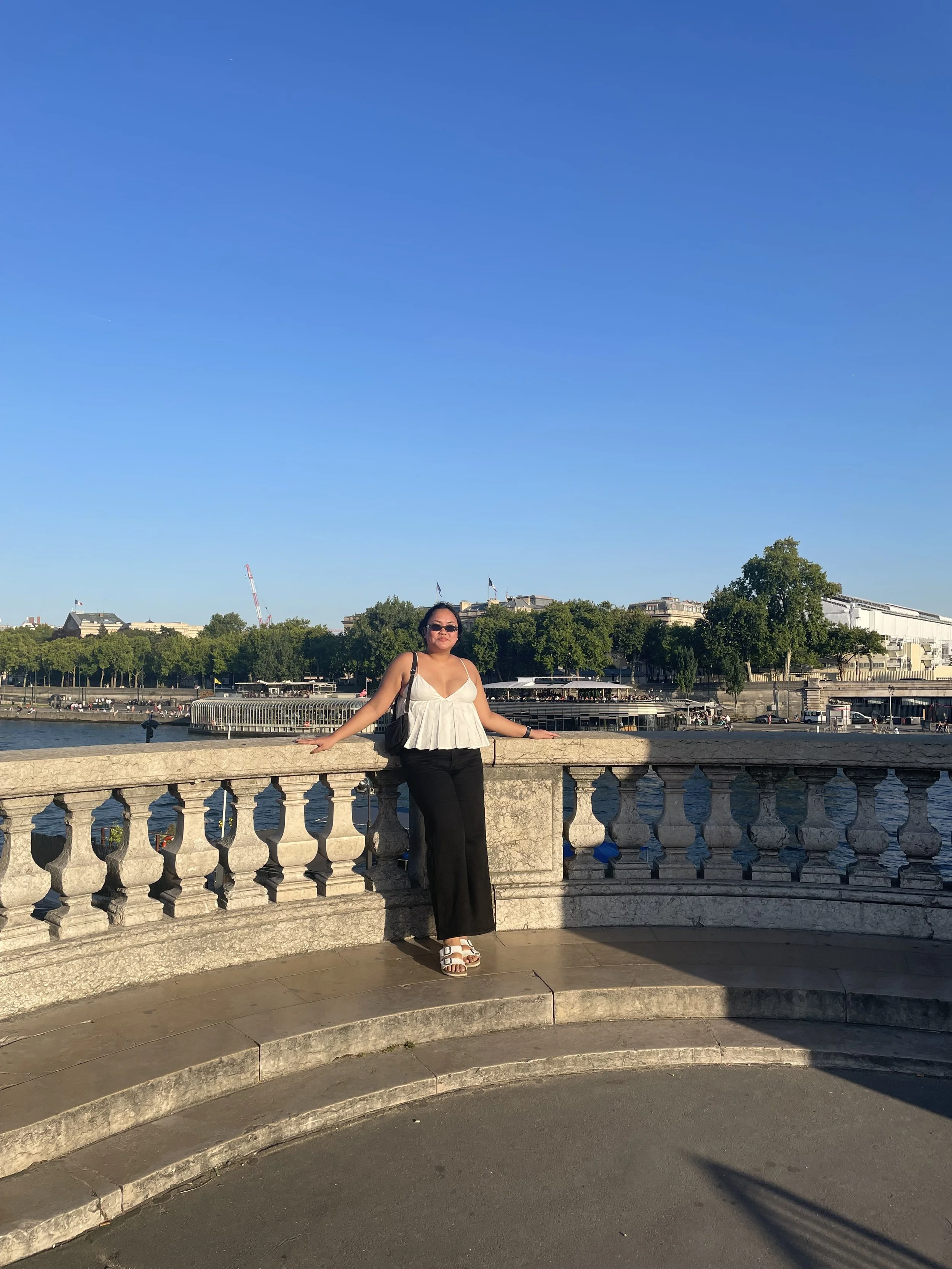 A woman in a white top and black pants standing by a stone railing near a river with trees and buildings in the background under a clear blue sky.