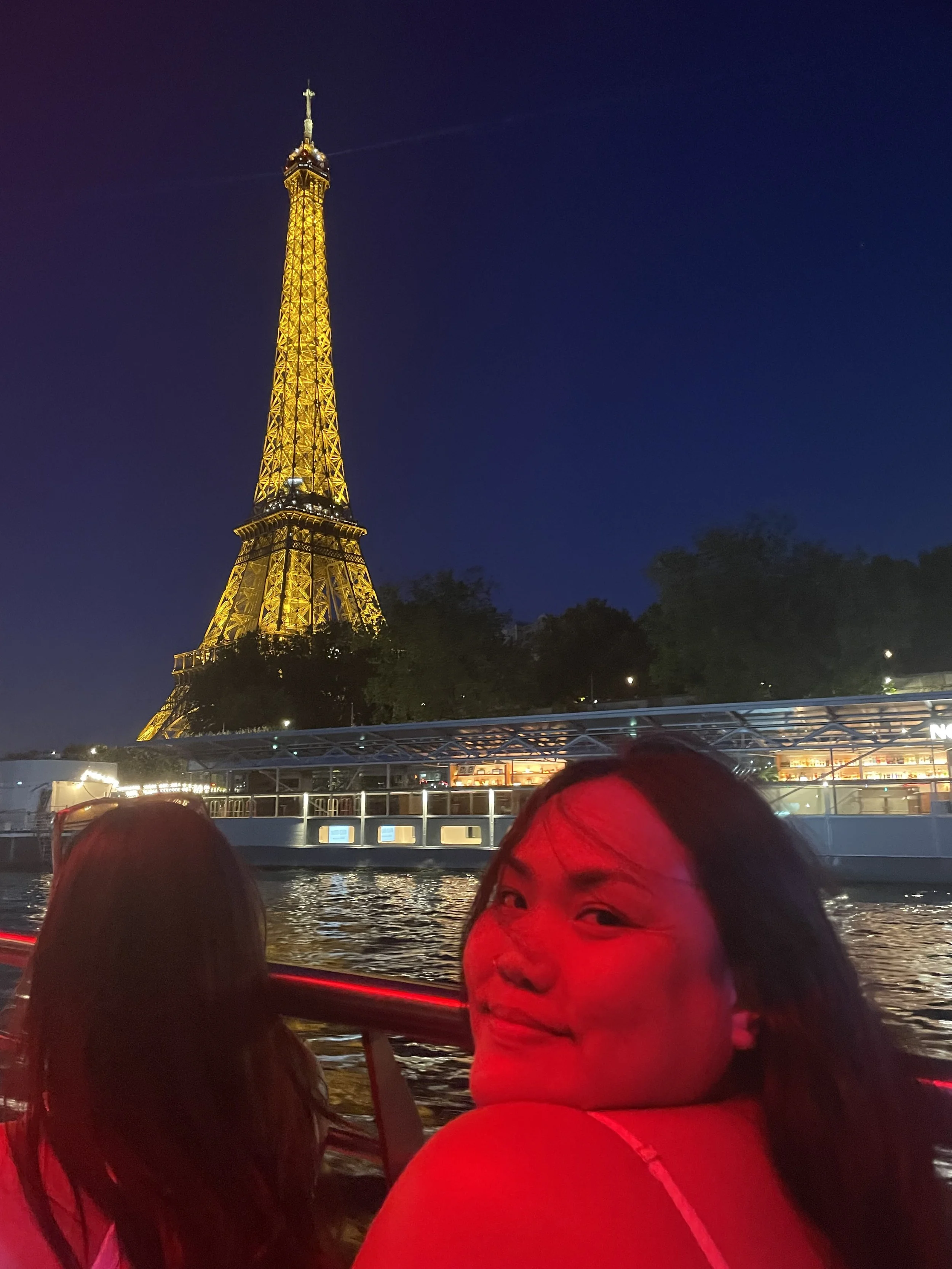Night view of the Eiffel Tower illuminated in gold, with two women in the foreground on a boat, one smiling, on the river in Paris