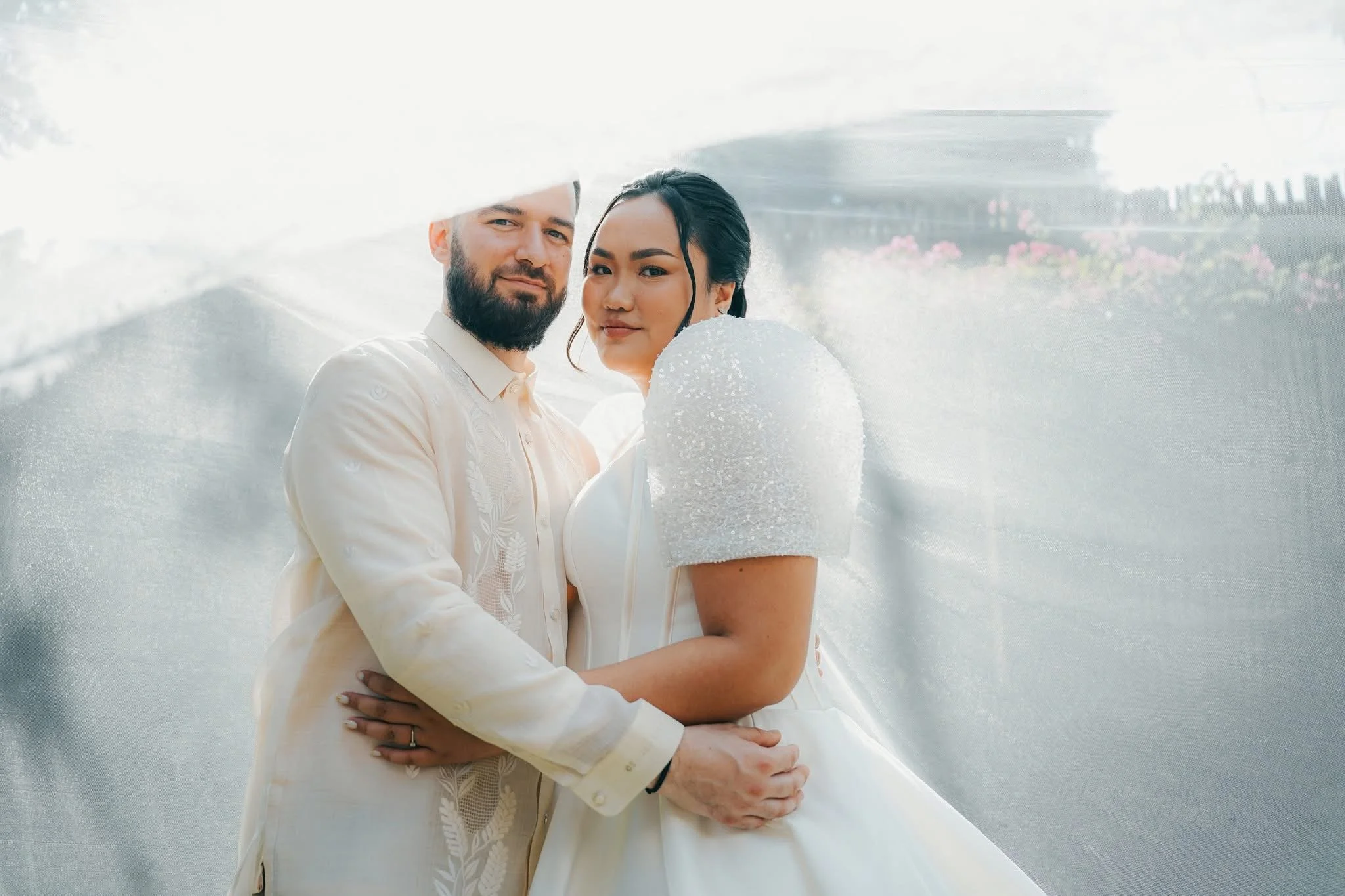 A couple dressed in white, standing close outdoors, with a modern building and pink flowers in the background.