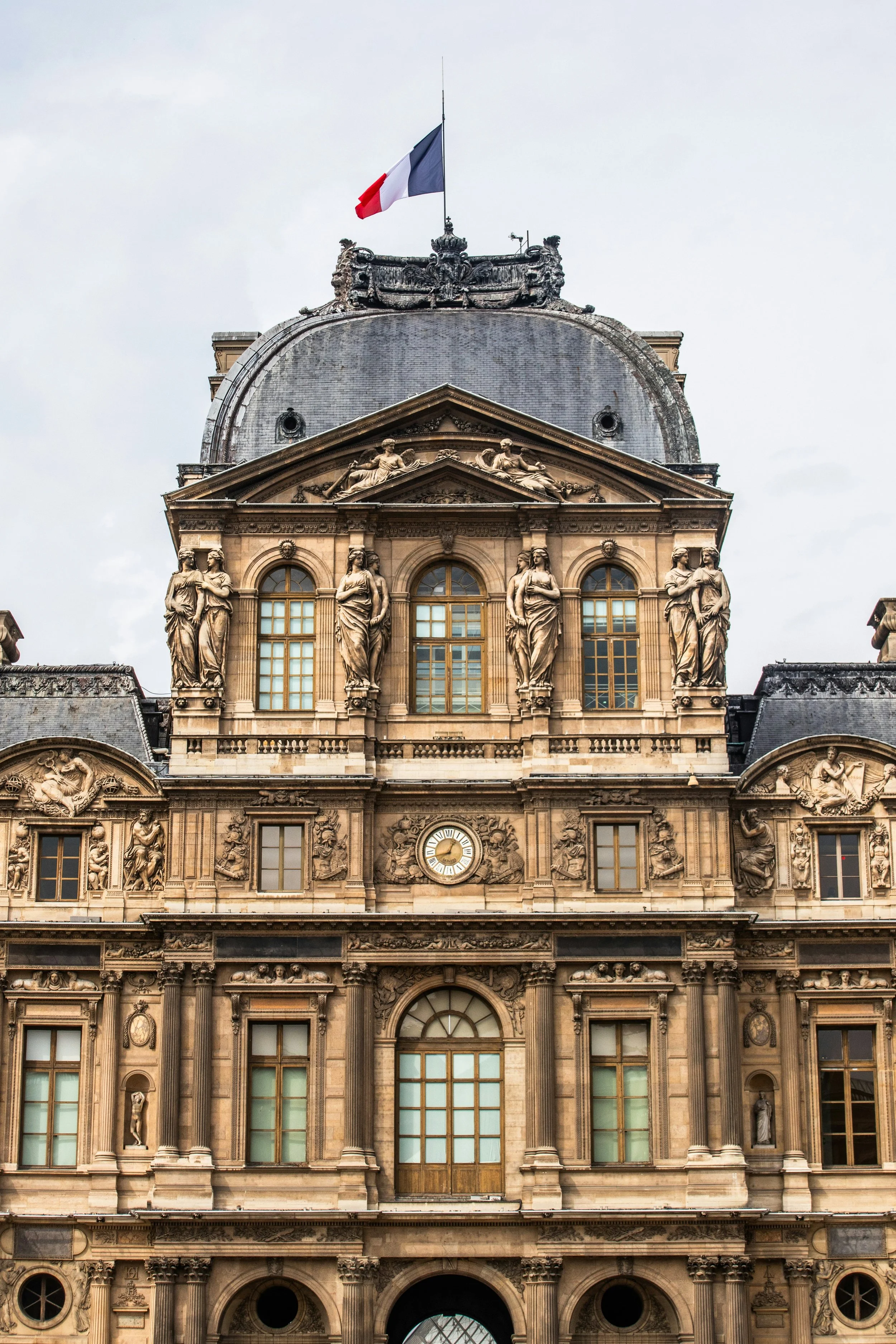 Front view of a historic building with ornate sculptures and details, topped with a French flag.