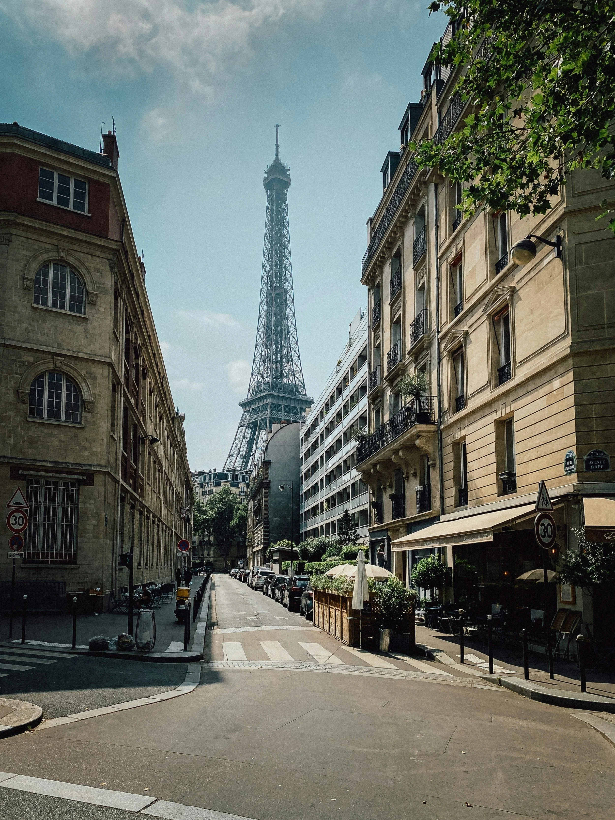 A Paris street scene with traditional Parisian architecture, parked cars, and a view of the Eiffel Tower in the background under a partly cloudy sky.