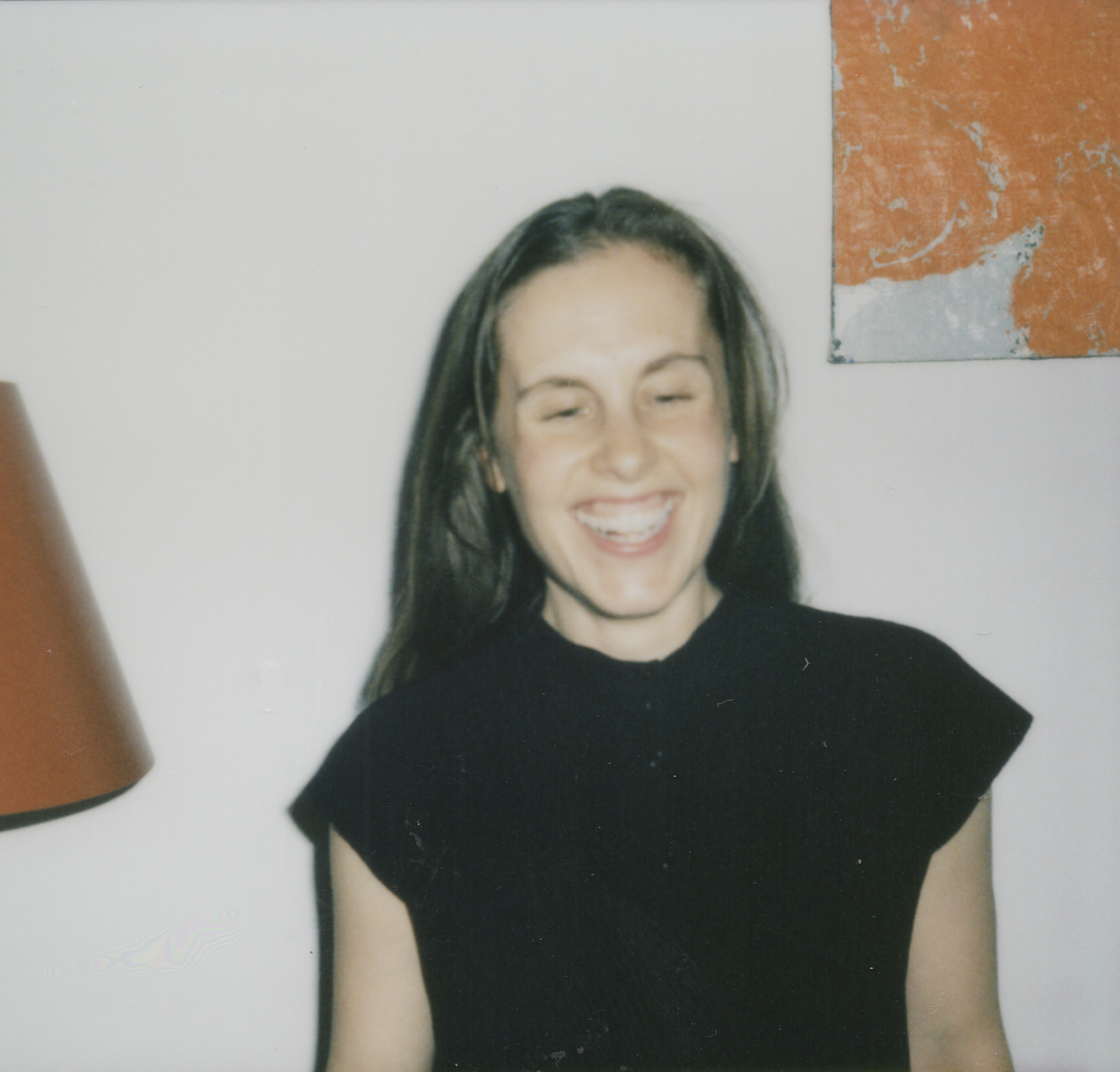 A woman with long dark hair smiling and laughing, wearing a black top, standing in front of a white wall with an orange and gray abstract painting and an orange lampshade partially visible on the left.
