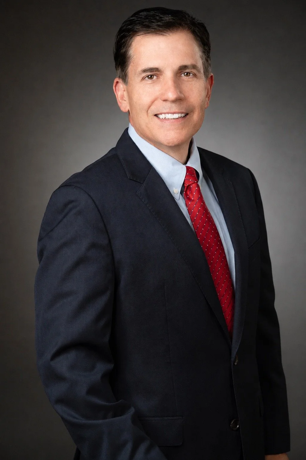 A man in a dark suit jacket, light blue shirt, and red patterned tie standing against a gray background, smiling at the camera.