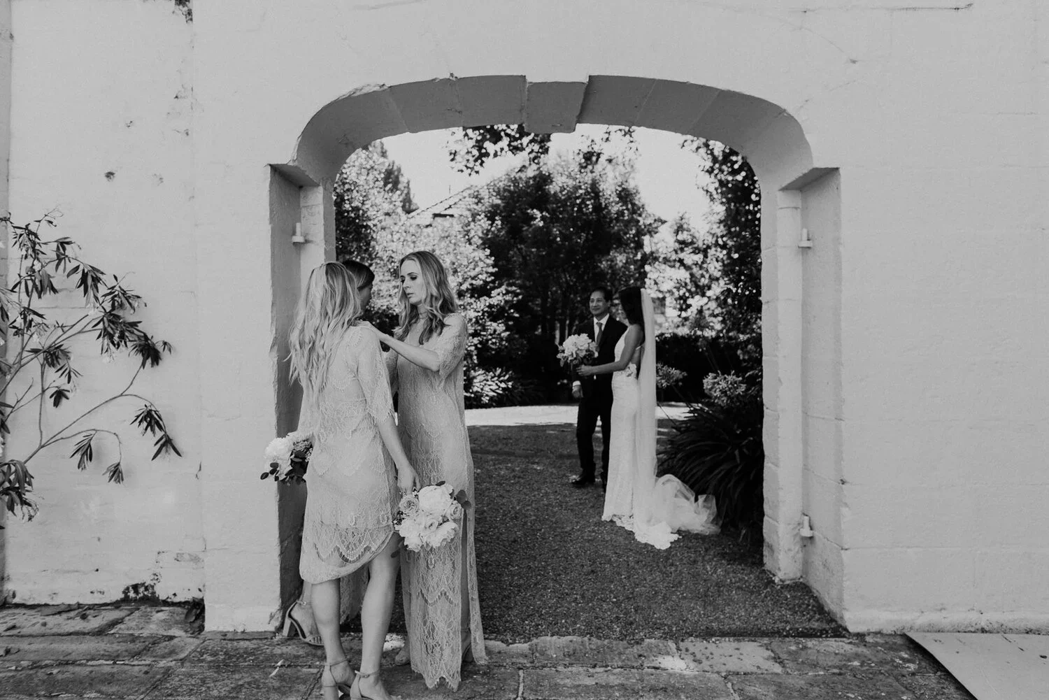 Black and white photo of a wedding scene with two women standing under an arch, with a bride and groom in the background holding bouquets of flowers.