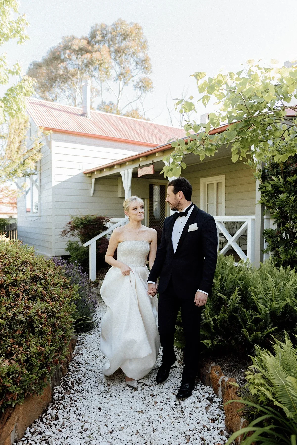 A bride in a strapless wedding gown holding hands with a groom in a black tuxedo, walking in a garden pathway with lush greenery and white gravel, in front of a white house with a red roof.