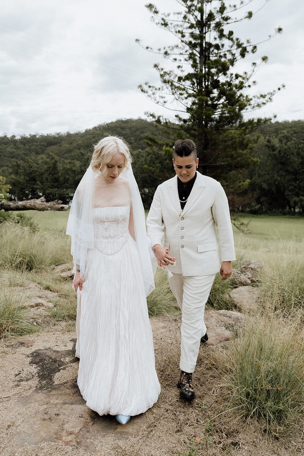 Two people, a brides on their wedding day with blonde hair and a brideswoman with dark hair, walk hand in hand outdoors in a grassy area with trees and hills in the background, dressed in wedding attire.