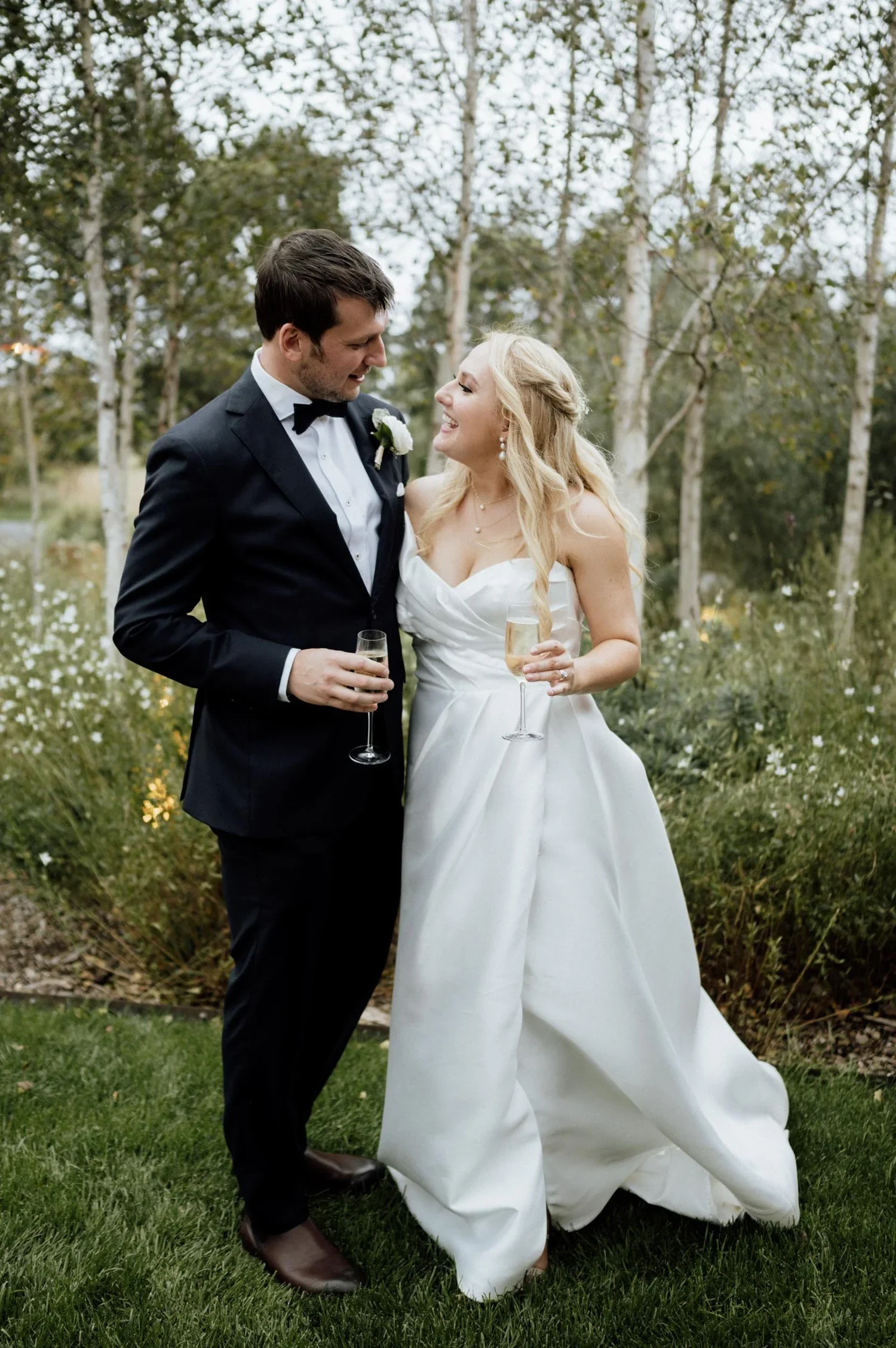 A bride and groom smiling and holding glasses of champagne outdoors in a natural setting with trees and grass.