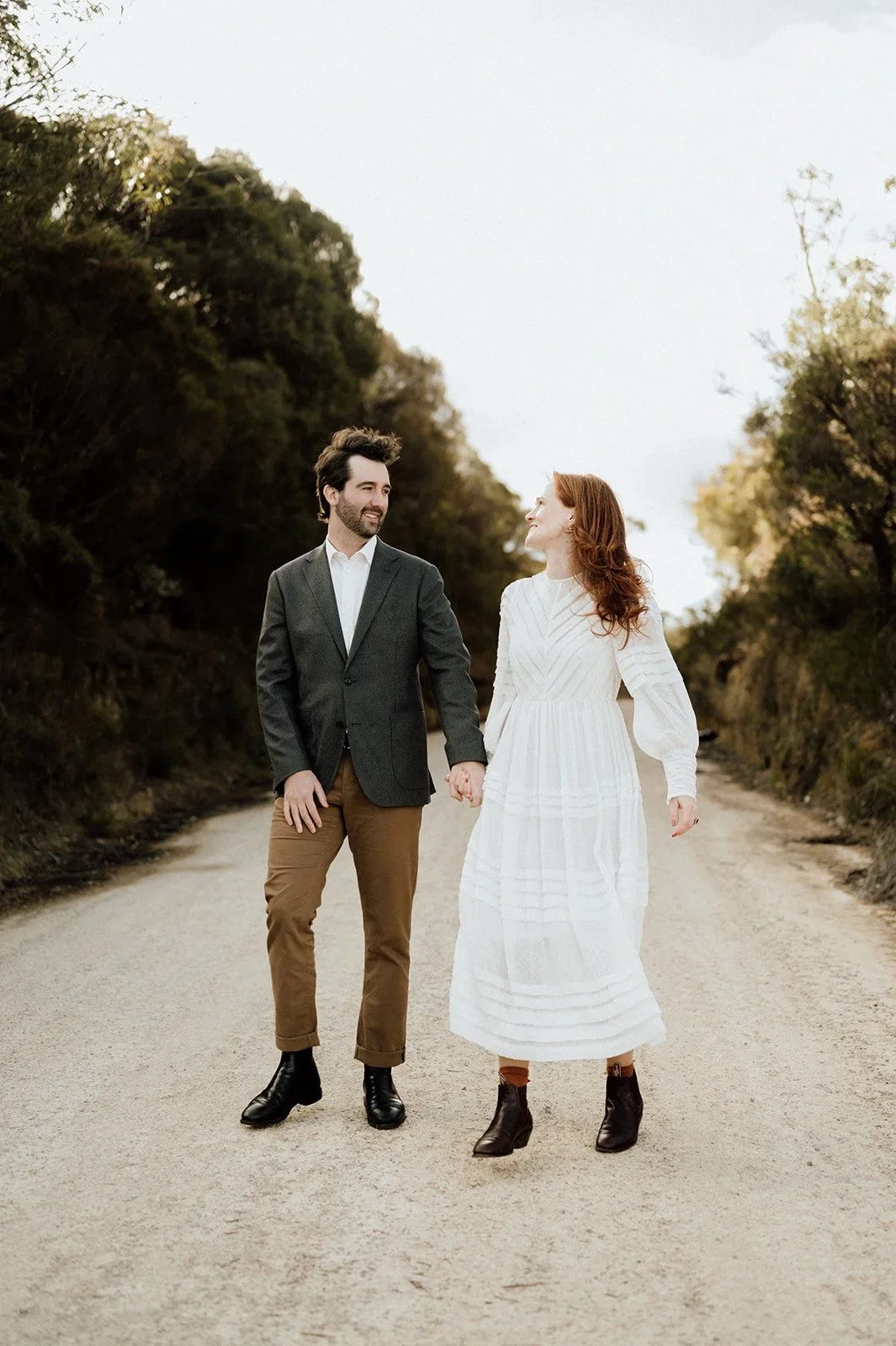 A couple holding hands and walking on a dirt road in a natural outdoor setting, with trees on both sides, during daytime.