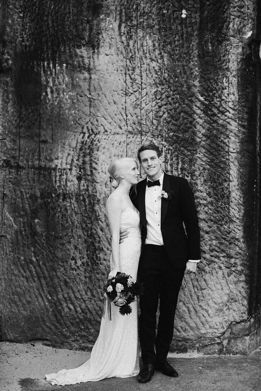 Black and white photo of a bride and groom standing close together, smiling, in front of a large textured rock wall. The bride is wearing a long wedding dress and holding a bouquet. The groom is wearing a tuxedo with a bow tie.