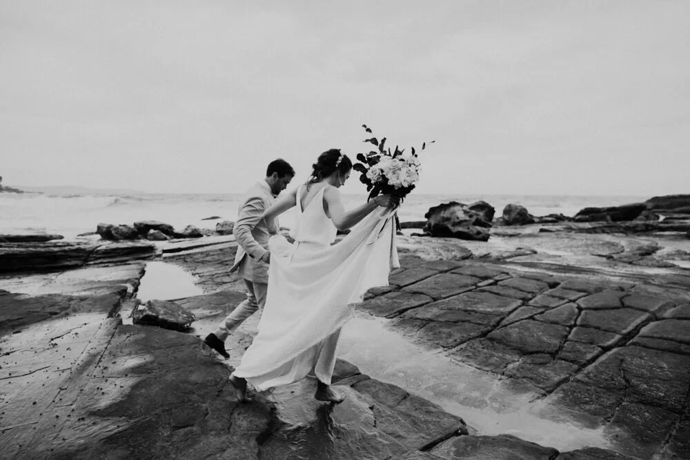 A couple in wedding attire walking barefoot on rocky beach, with the woman carrying a bouquet of flowers and the man walking behind her.