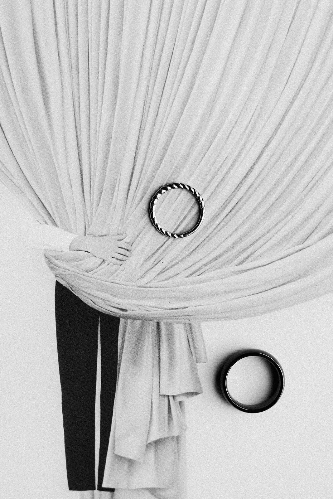 Black and white photo of a person holding back a curtain with a ring on it, showing their arm and part of their leg, with another ring on the floor.