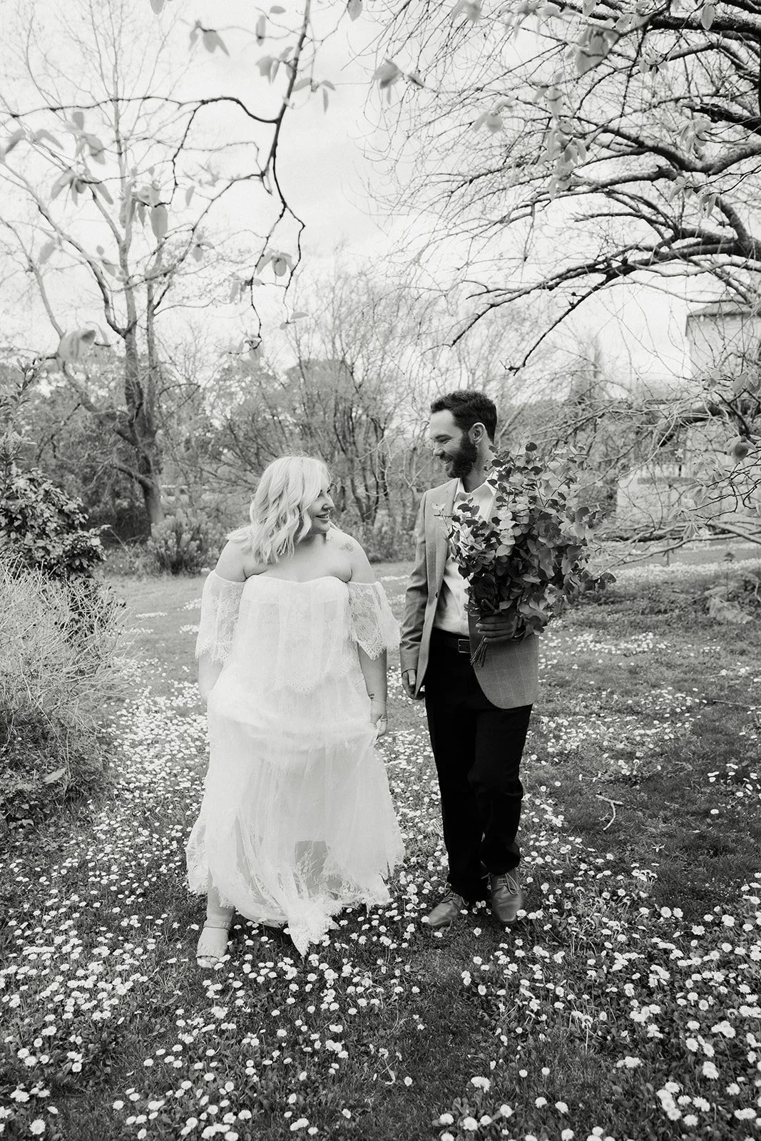 A couple walking in a garden, the woman in a white dress, the man holding a bouquet of flowers, both smiling and looking at each other, surrounded by trees and flowers.