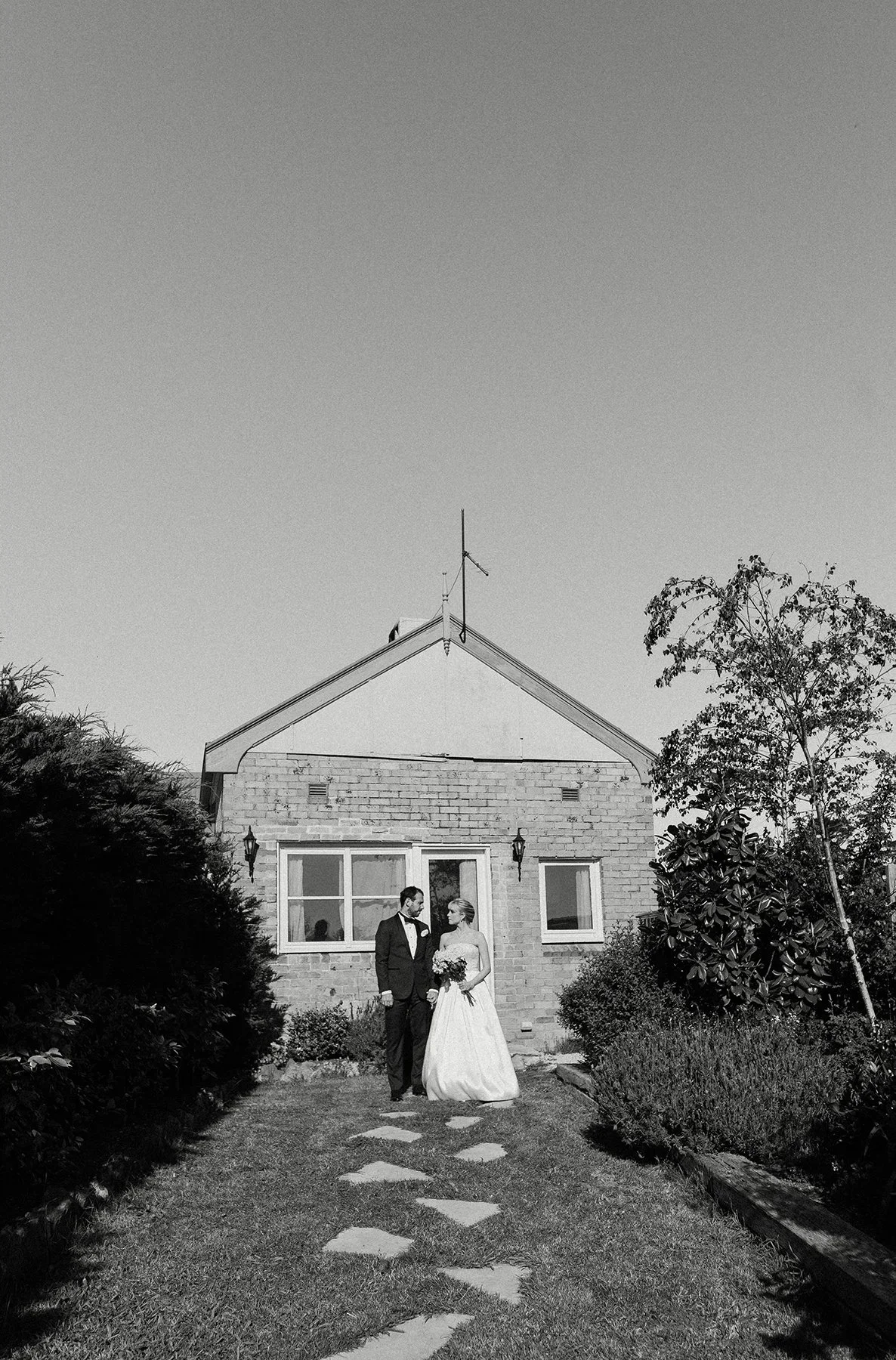 A wedding couple standing in front of a house with trees and bushes on either side, holding hands and looking at each other, in black and white.