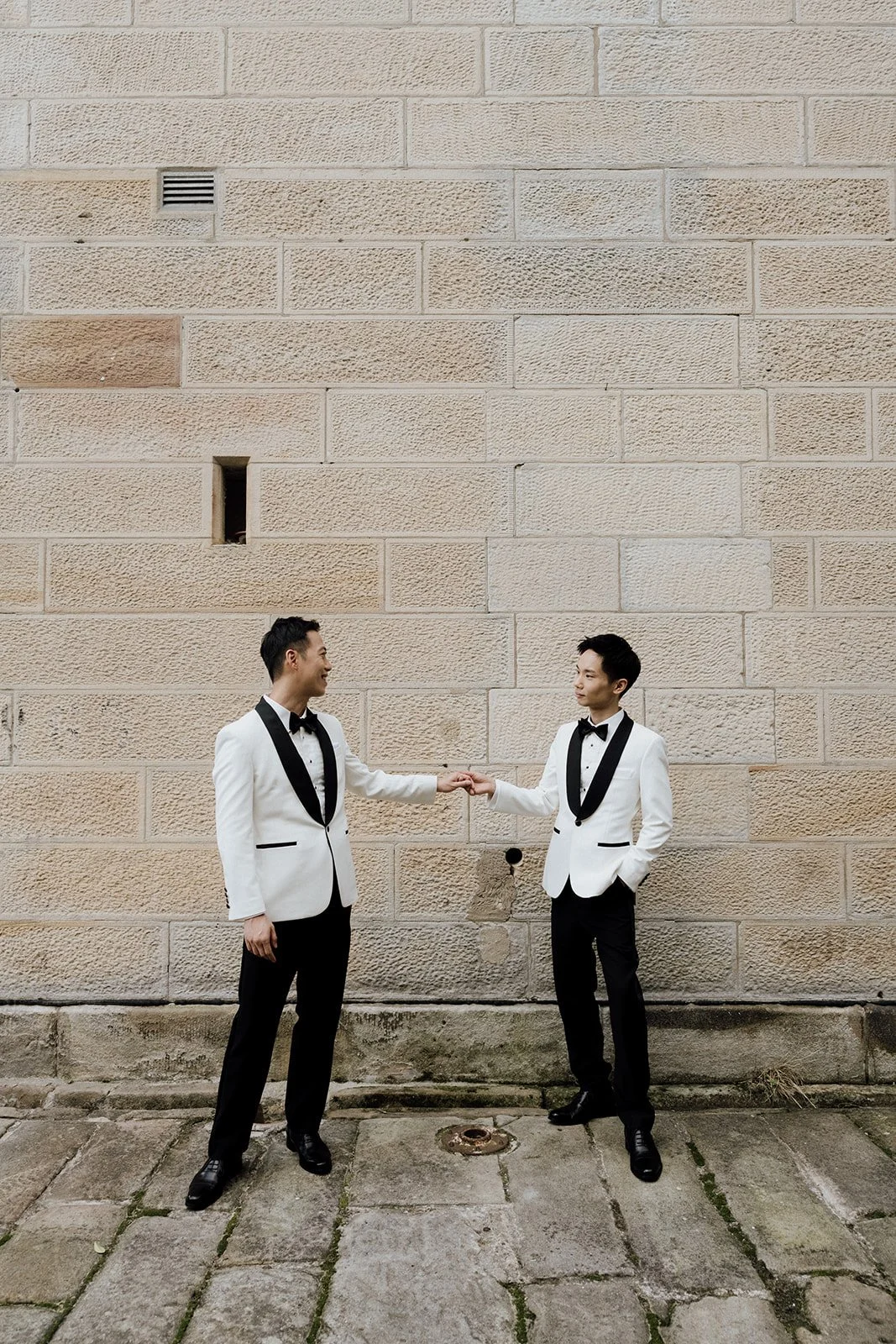 Two men in tuxedos with white jackets and black bow ties are standing and shaking hands in front of a beige brick wall, sydney inclusive wedding photographer.