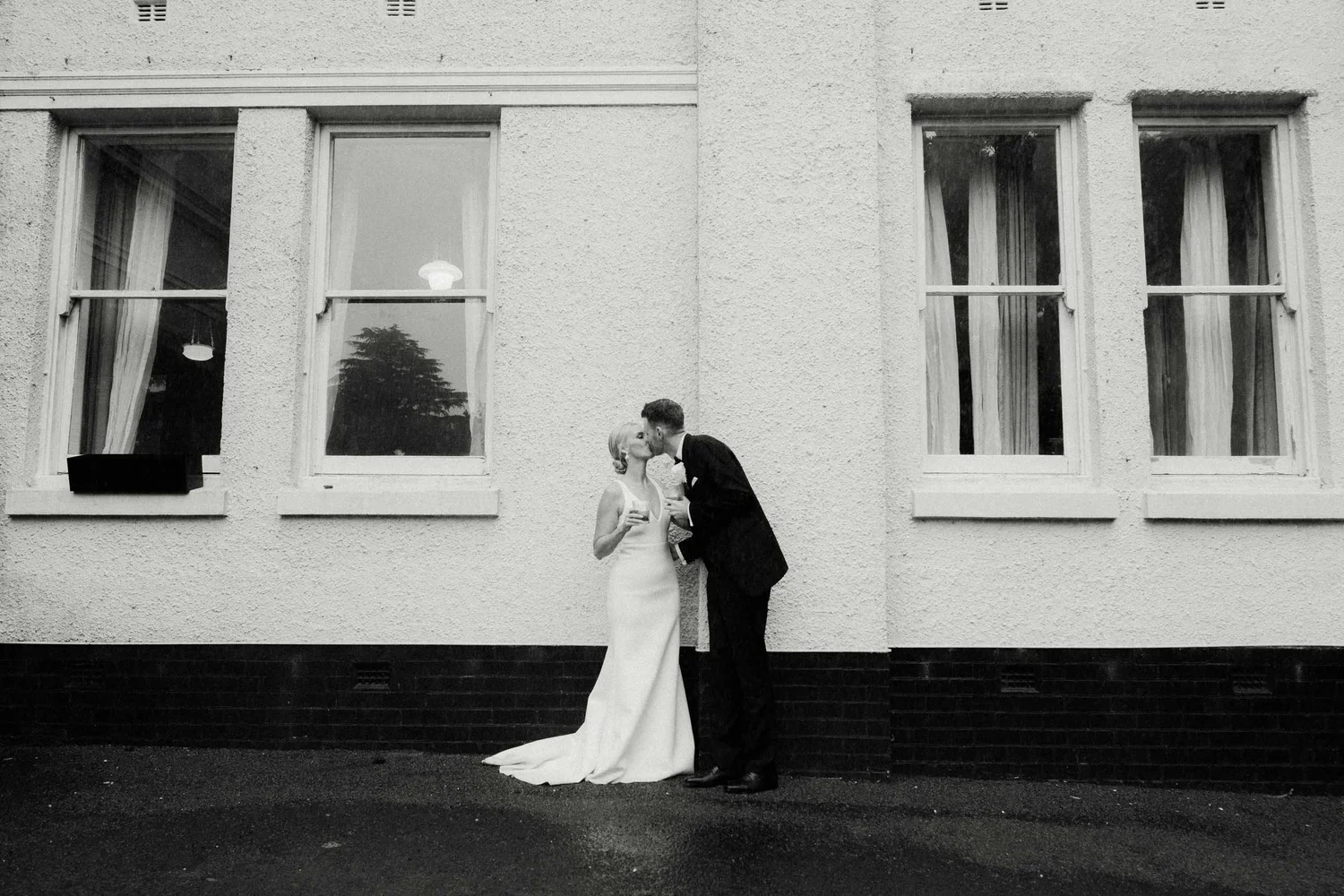 Black and white photo of a newlywed couple kissing outside a building with four large windows, the bride in a white gown and holding a drink, the groom in a suit.