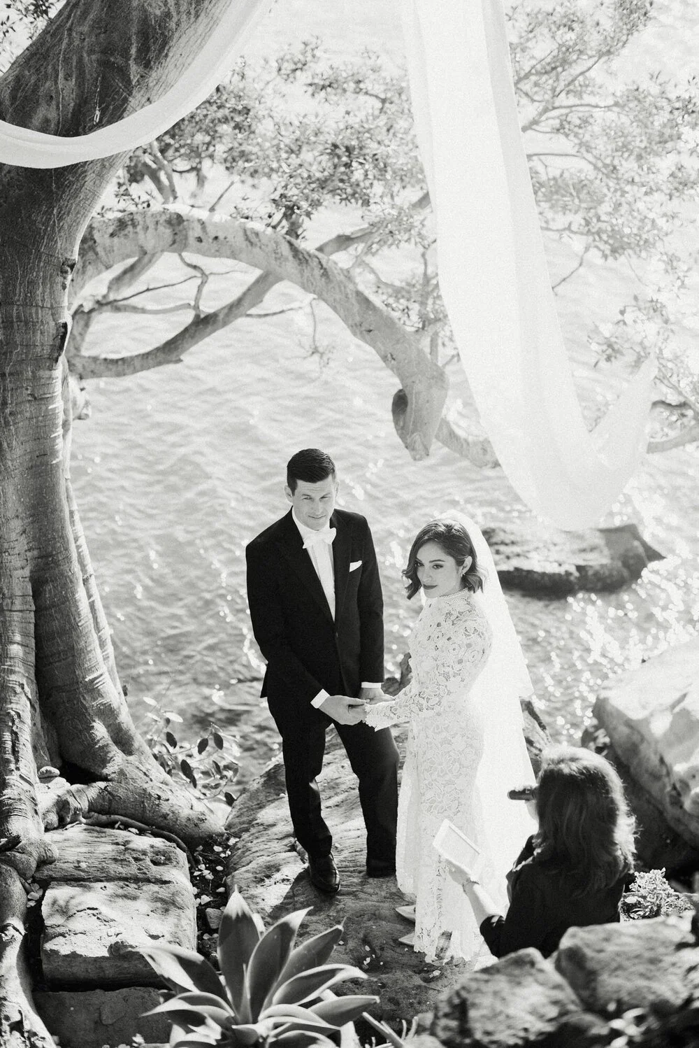 A wedding ceremony taking place outdoors near the water, with a bride and groom holding hands and facing each other, a woman officiating, and a large tree in the background.
