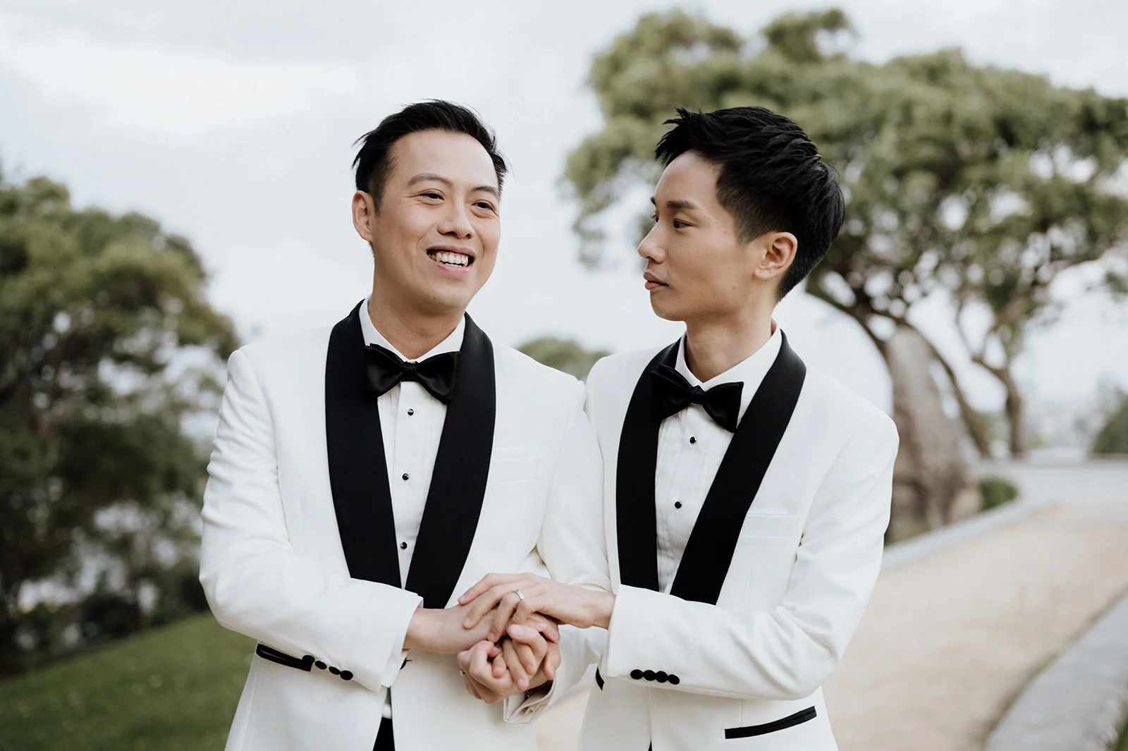 Two men in white tuxedos with black lapels and bow ties, holding hands and smiling outdoors with trees in the background.