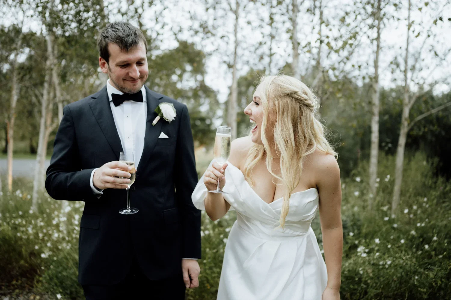 A bride and groom in wedding attire celebrating outdoors with glasses of champagne, smiling and laughing.