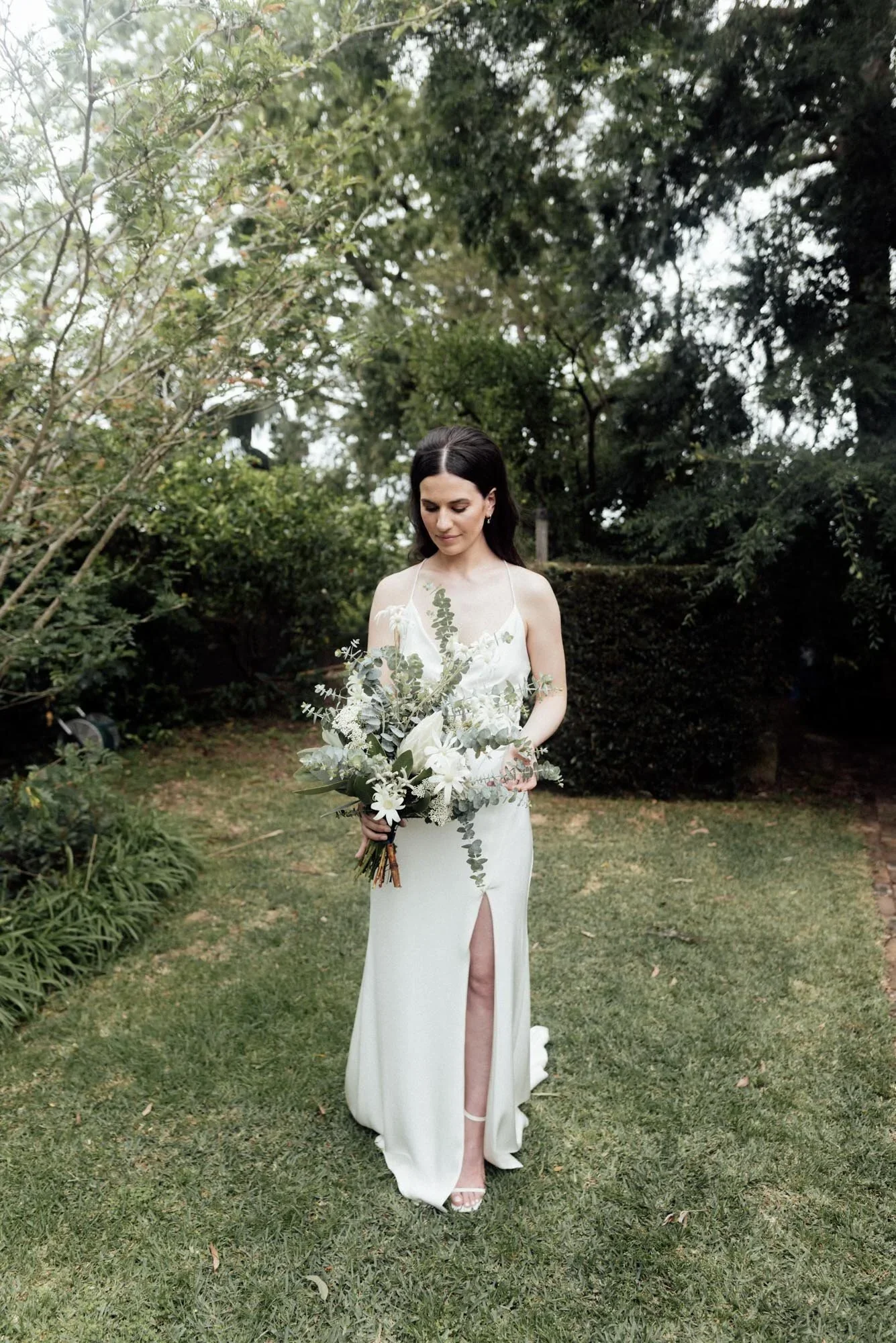 A woman in a white dress holding a bouquet of white flowers stands on a grassy lawn surrounded by trees and bushes.