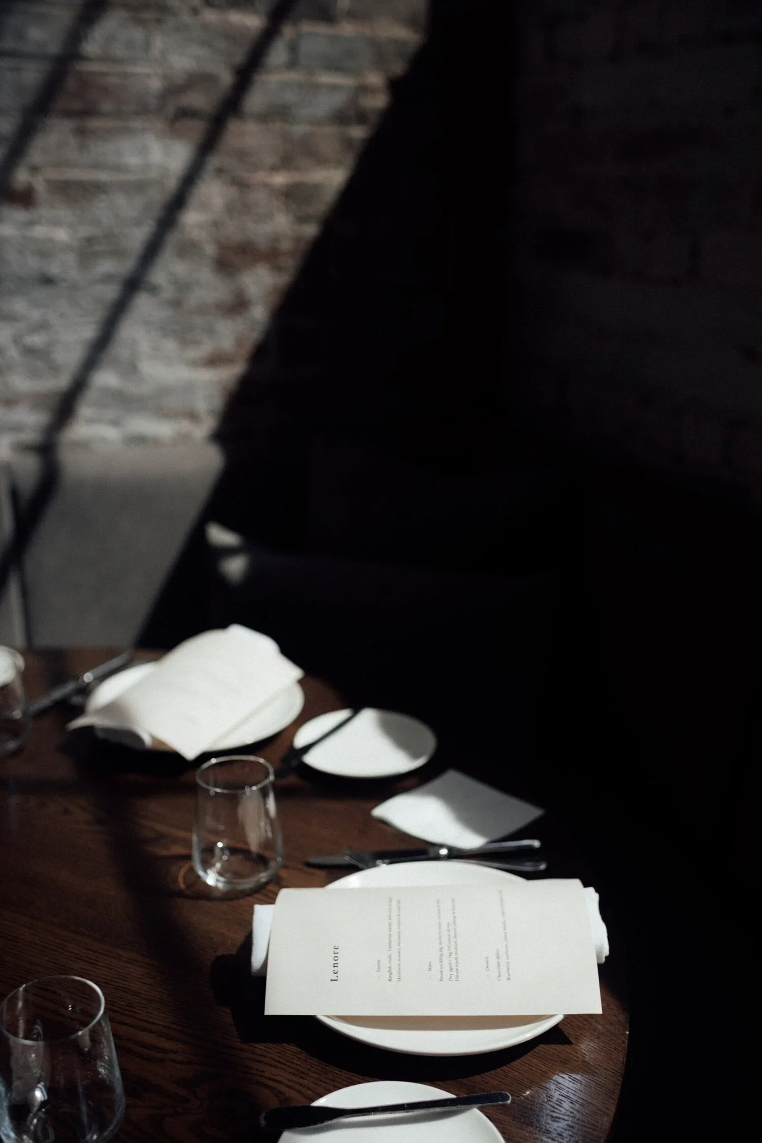 A restaurant table set with white plates, clear drinking glasses, silverware, napkins, and a menu, with a dark wood surface and brick wall in the background.