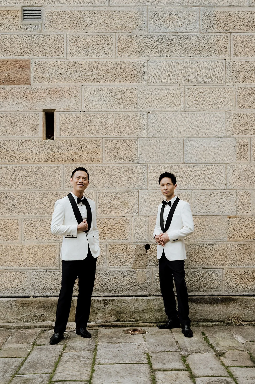 Two men in tuxedos standing against a beige brick wall, smiling and holding their lapels, same sex wedding at gunners barracks in sydney.